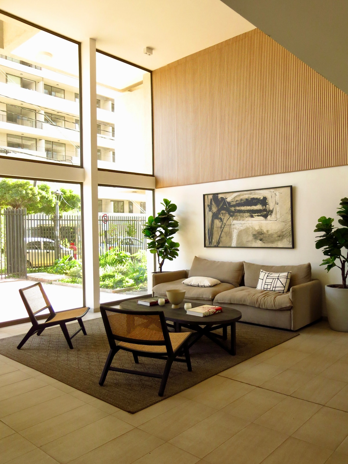 A modern lobby area is presented, featuring large windows that allow natural light to fill the space. A beige sofa is complemented by two wooden chairs and a coffee table. The room displays potted plants and a wall art piece, enhancing the inviting environment.