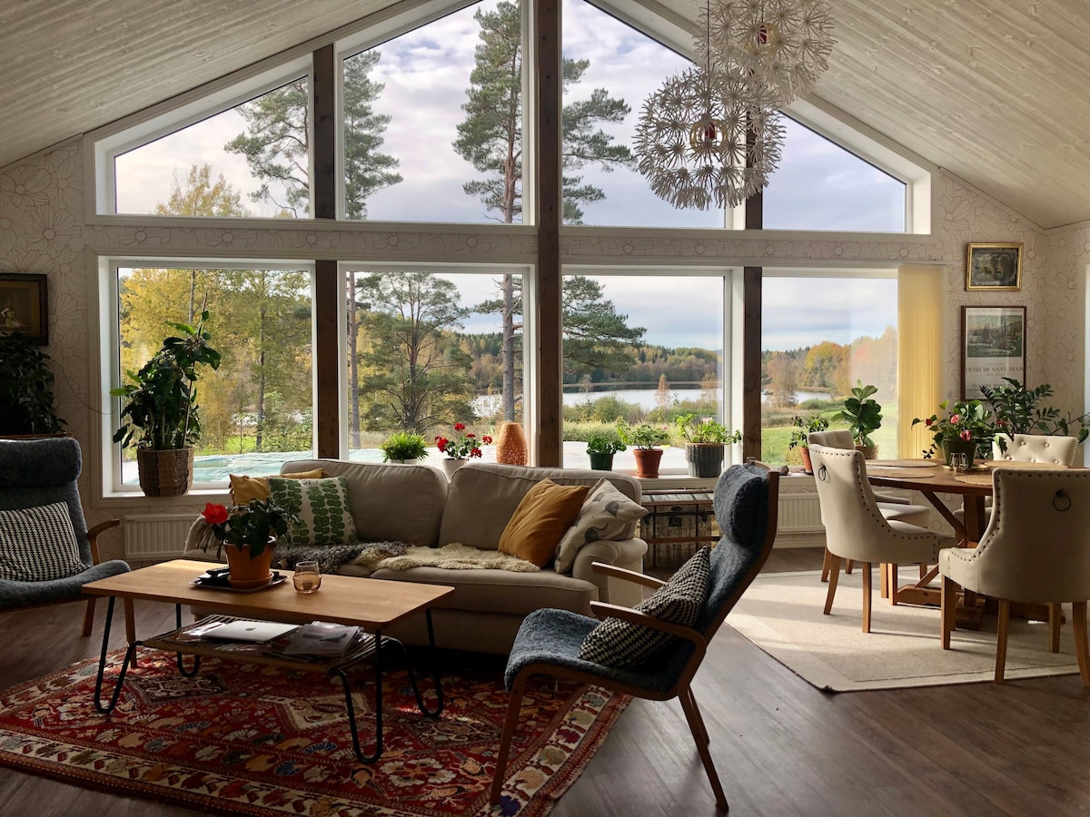 A spacious living area is illuminated by large windows offering lake views. Comfortable seating arrangements include a sofa and armchairs. A wooden coffee table rests on a patterned rug, while a dining area accommodates multiple chairs around a table. Potted plants add greenery to the scene.
