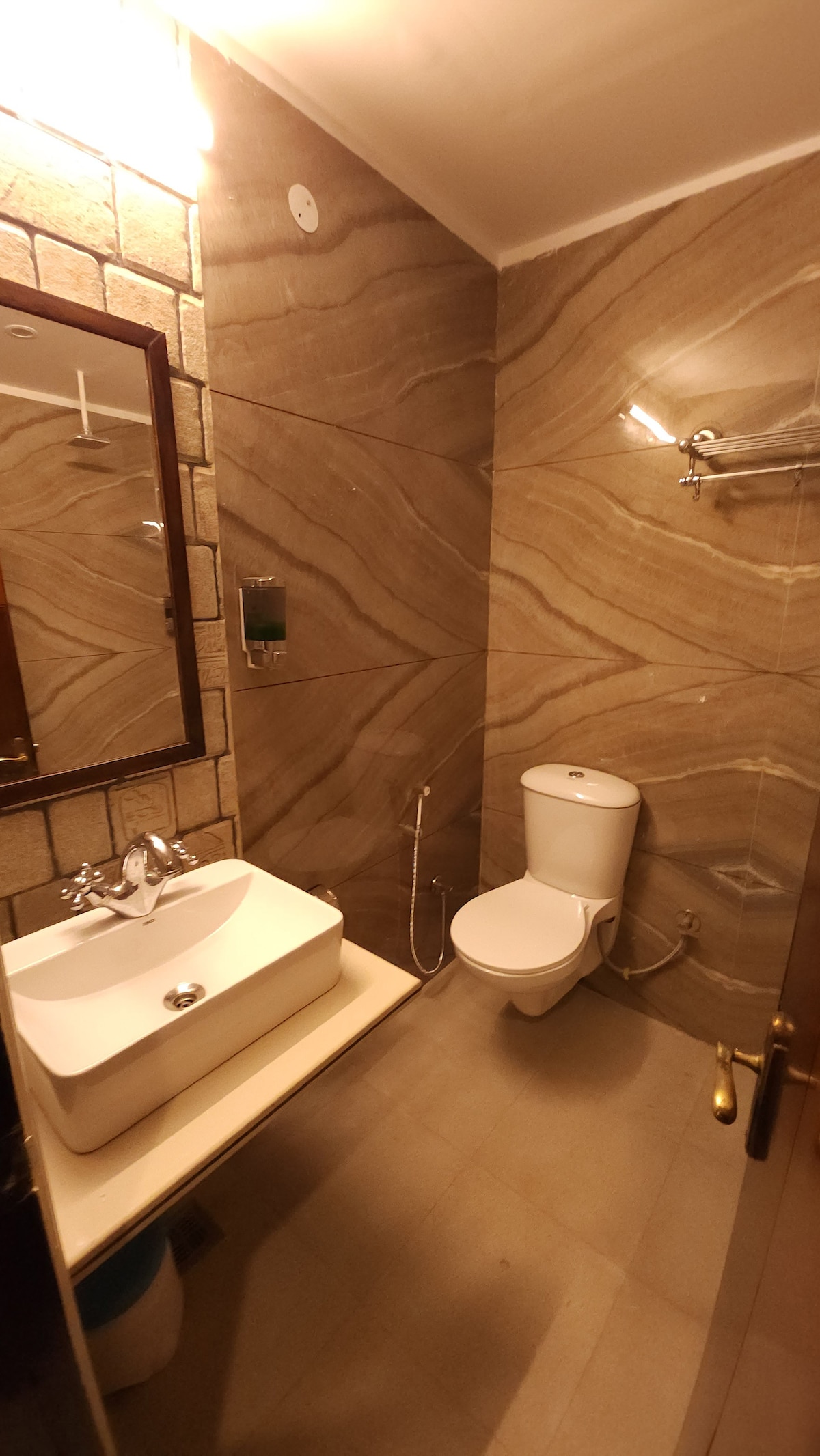 A modern bathroom features beige and brown stone walls, providing a sophisticated look. A sleek white sink is positioned on a light countertop, accompanied by a large mirror above. The toilet is located beside a built-in wall shelf, adding functional space.