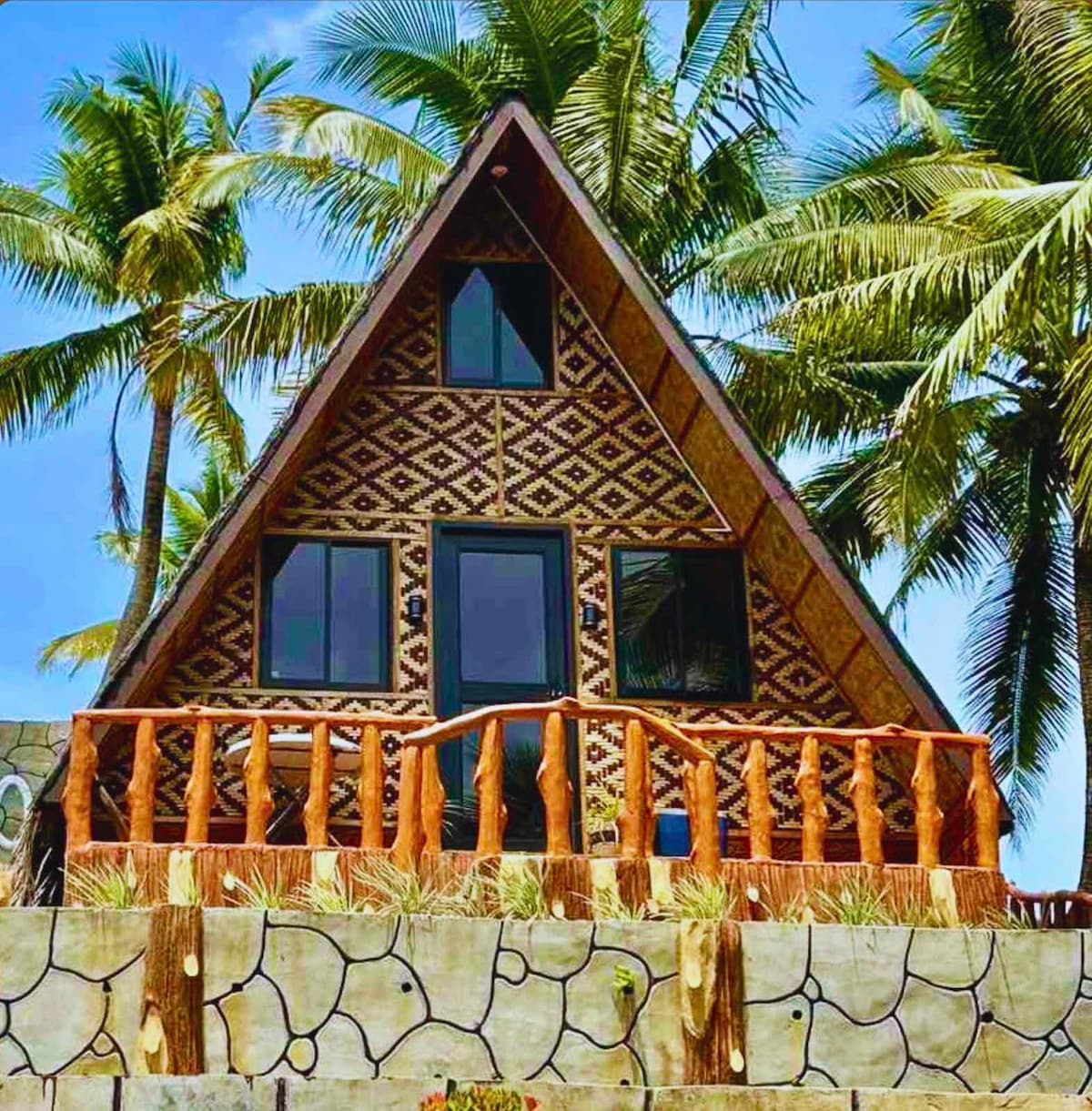 The exterior of an A-frame house is showcased, featuring intricate patterns on the façade. Tall palm trees frame the structure, while a wooden railing surrounds the front porch, offering a warm and natural setting in the inviting sunshine.