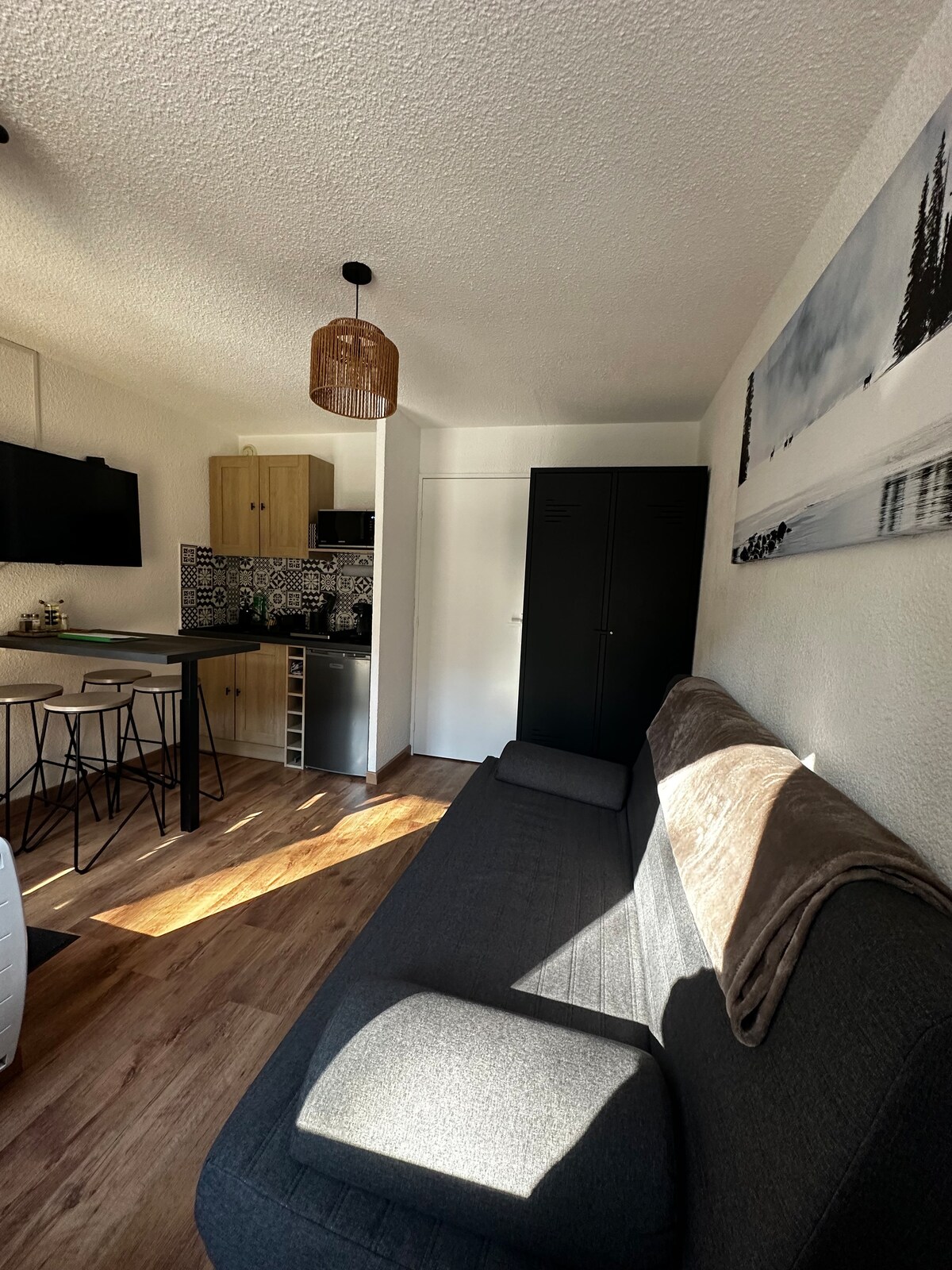 A modern living area features a comfortable grey sofa and a cozy throw blanket. A kitchen area with wooden cabinetry and patterned backsplash is visible in the background, while a dining space with slender black stools is situated nearby. Natural light enters through a window, enhancing the inviting space.