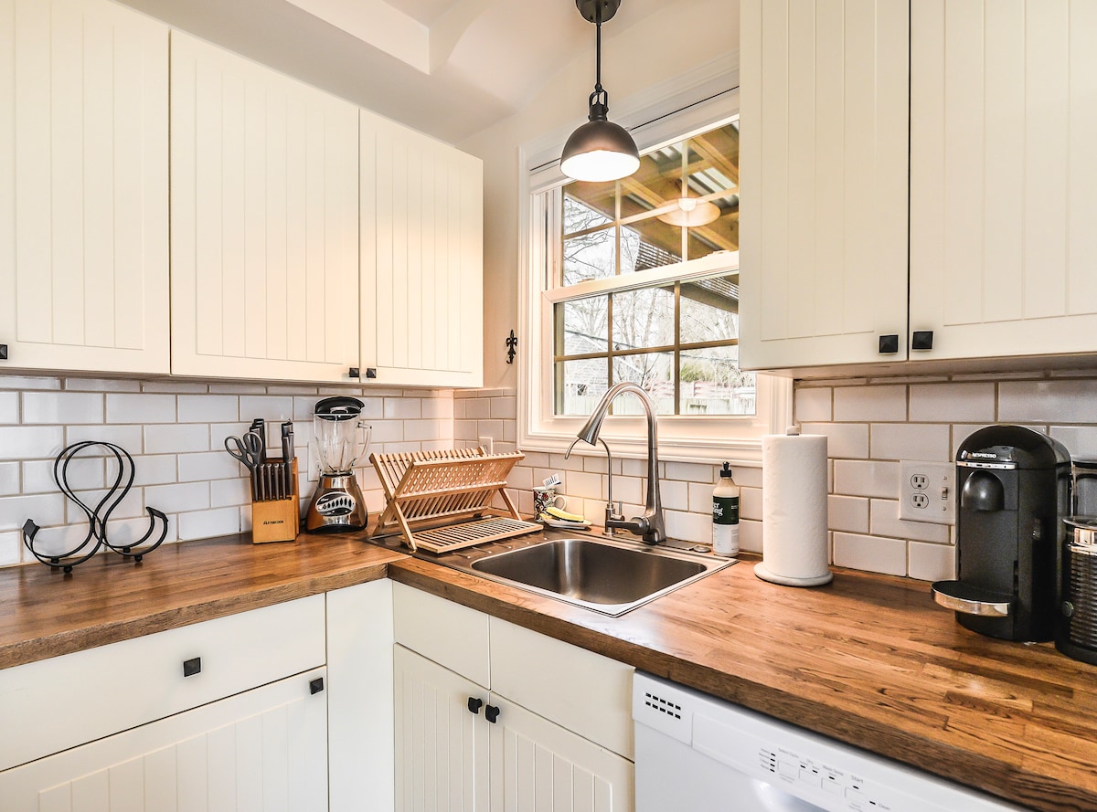 A well-equipped kitchen features wooden countertops and white cabinetry. A window allows natural light to illuminate the space, complemented by a sink and modern appliances, including a dishwasher and a coffee machine. Kitchen utensils and a drying rack are neatly arranged for convenience.