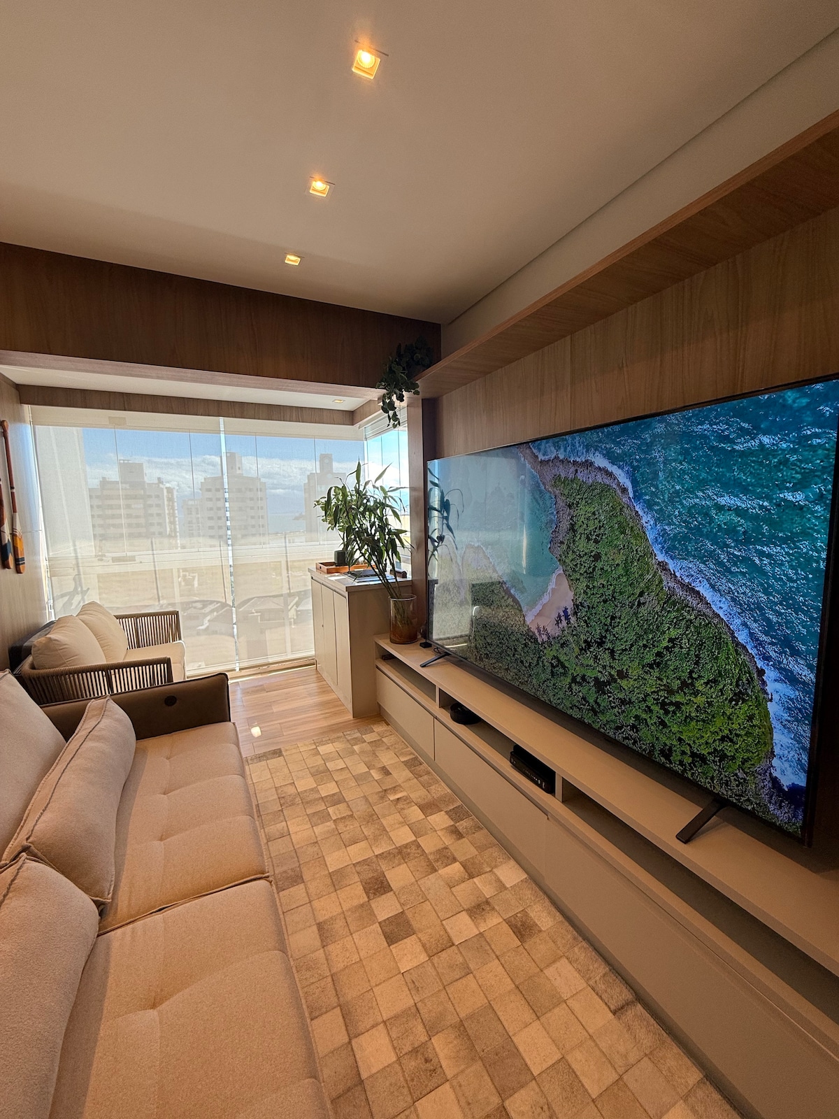 A modern living area is shown featuring a large television mounted on a wooden wall. A comfortable beige sofa is positioned across from the TV, with natural light filtering through a large glass window, providing a view of the city outside.