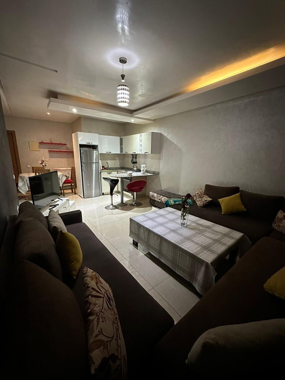 A cozy living area features modern furnishings with a large, L-shaped sofa adorned with decorative pillows. A small coffee table sits in the center, surrounded by natural light from the nearby window. The kitchenette is visible in the background, equipped with essential appliances.