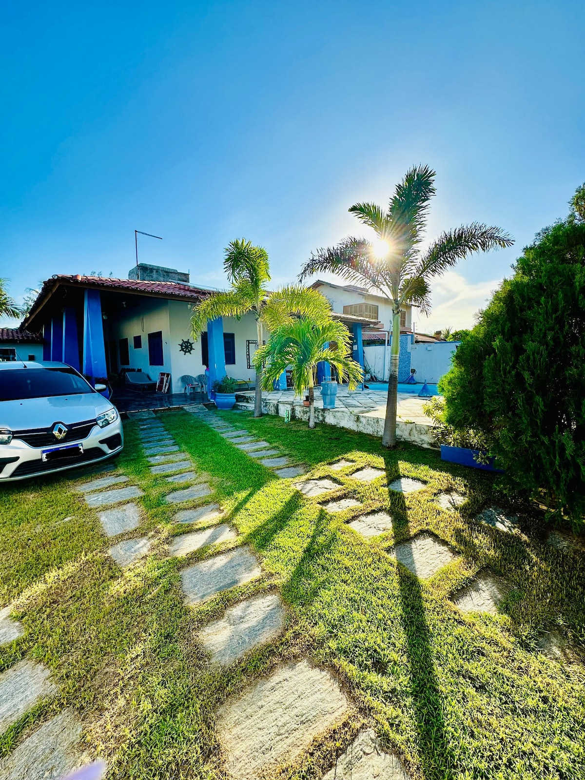The exterior of the house is presented, featuring a well-maintained garden with palm trees and a stone pathway leading to the entrance. A parked car is visible in front, with sunlight casting elongated shadows across the lush green grass.