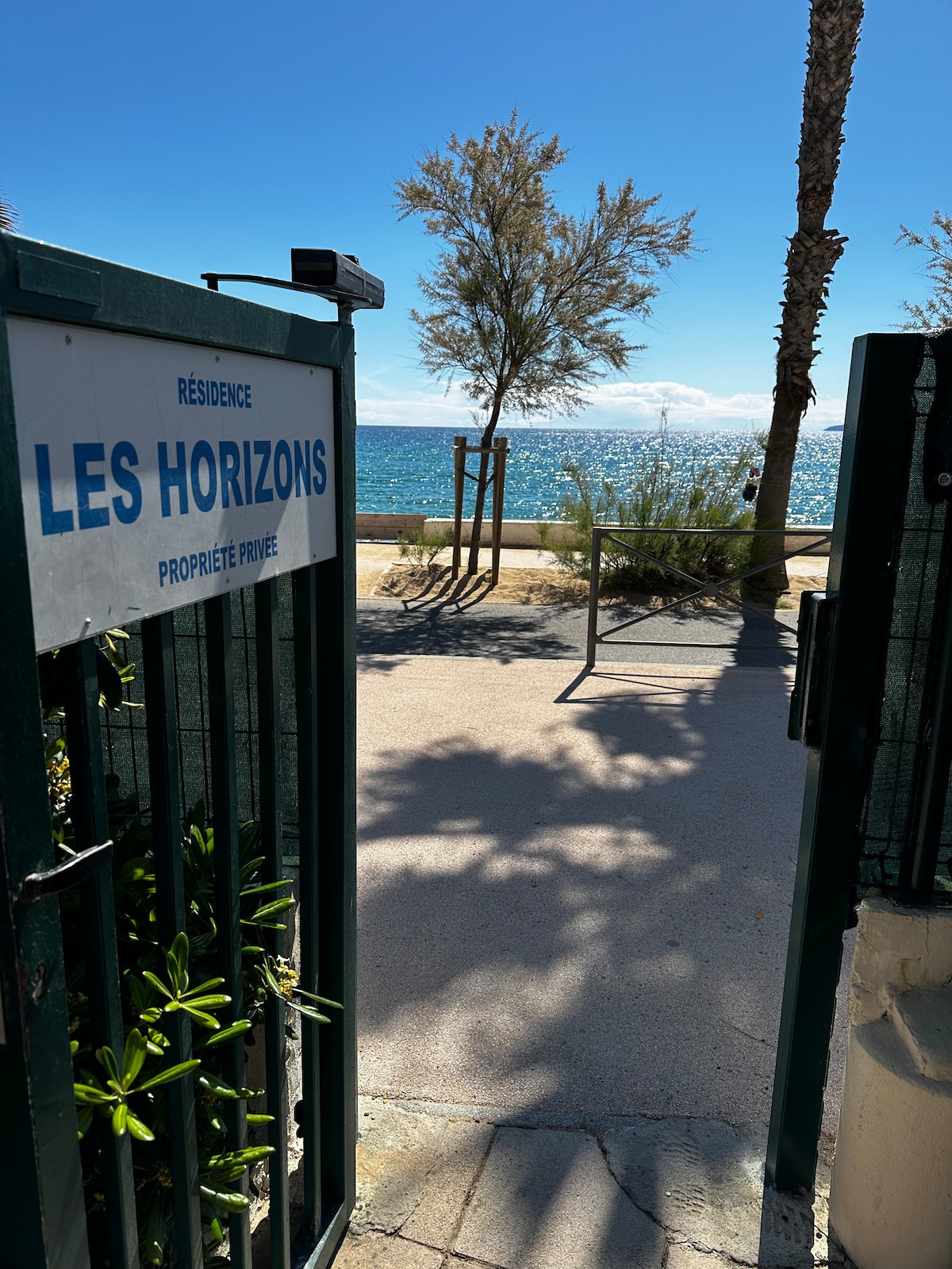 The entrance gate of Résidence Les Horizons opens to a scenic pathway leading to the beach. Lush greenery and palm trees are visible along the path, with the sparkling blue water of the Mediterranean Sea in the background under a clear sky.