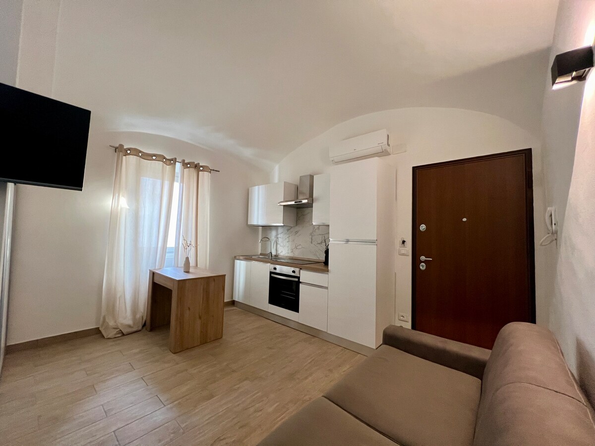 A modern kitchen area is visible, featuring a light wood dining table and sleek white cabinets. A comfortable beige sofa sits in the corner. Natural light filters through sheer curtains, enhancing the bright, airy feel of the space. A television is mounted on the wall.