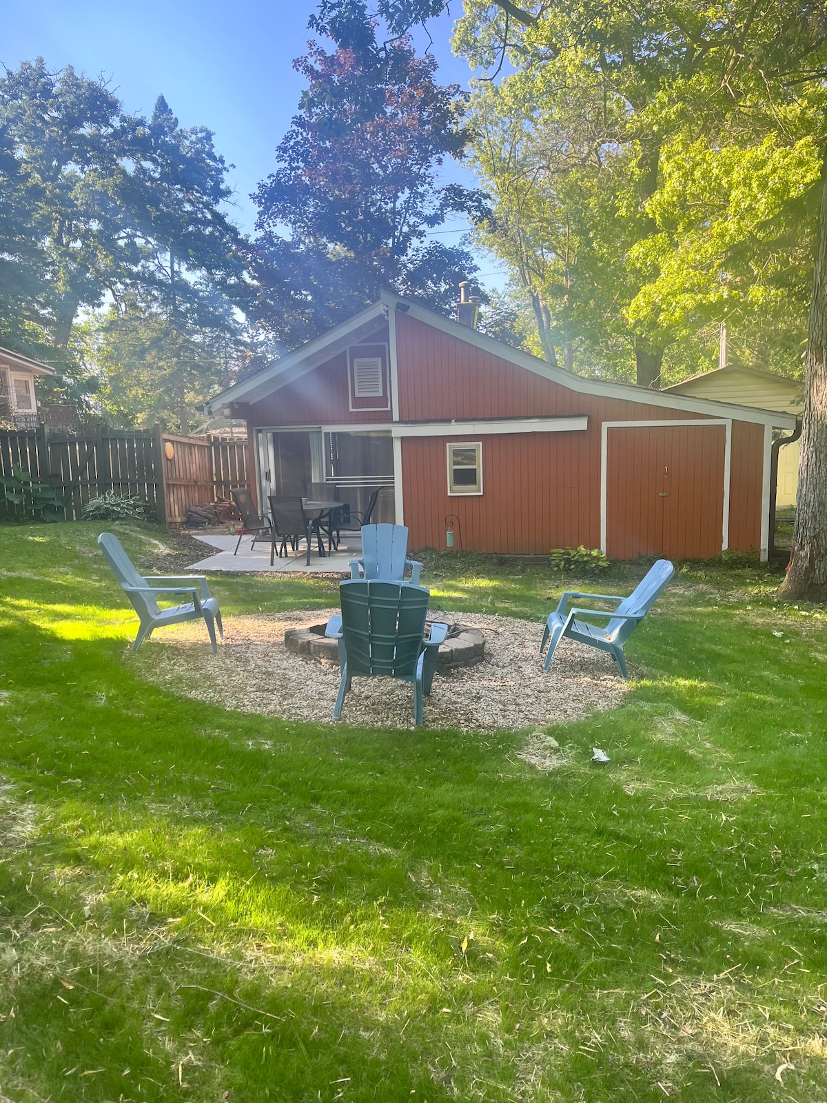 A spacious backyard features a fire pit surrounded by four blue Adirondack chairs, inviting relaxation. A small patio area is visible with additional seating, while a charming red bungalow stands in the background, framed by lush greenery and tall trees.