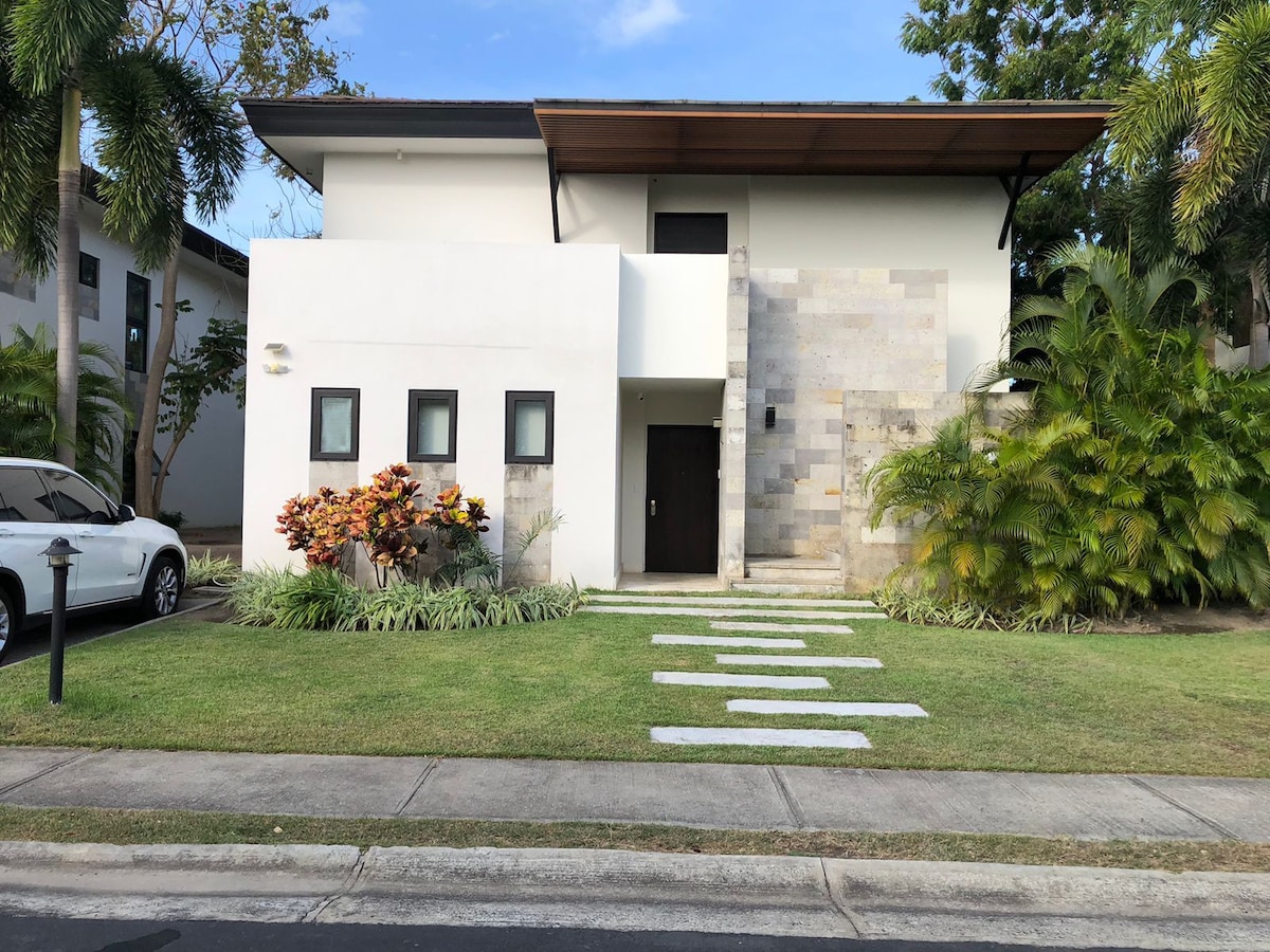 The two-story duplex features a minimalist design with a light-colored exterior. Stone accents adorn the entrance, complemented by a landscaped yard with greenery and colorful plants. A stone pathway leads to the front door, creating an inviting approach.