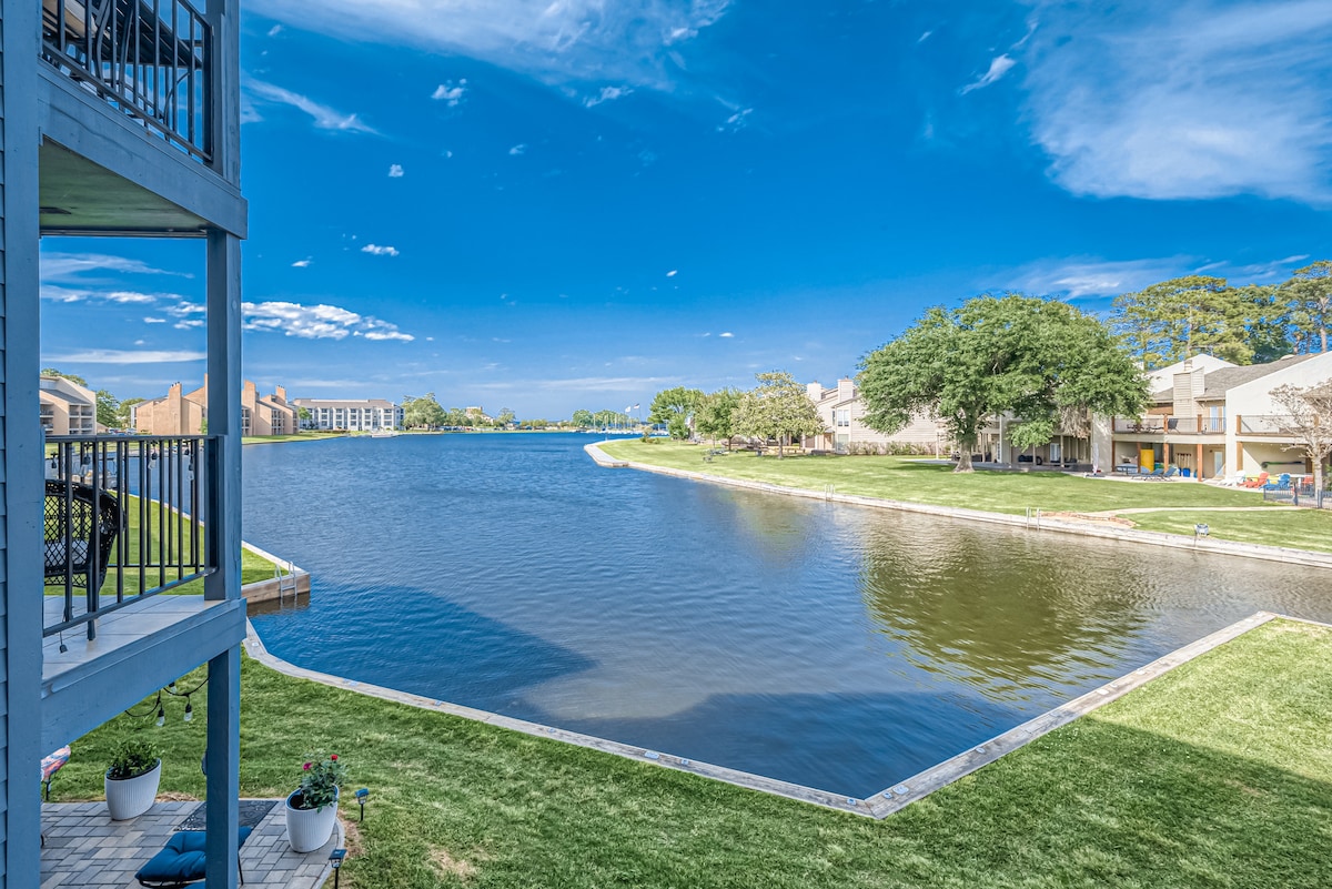 A scenic view of a calm waterway is captured, bordered by manicured green grass and lined with residential buildings. The clear blue sky reflects off the water, creating a serene backdrop ideal for relaxation. The image showcases the inviting outdoor space of the condo.