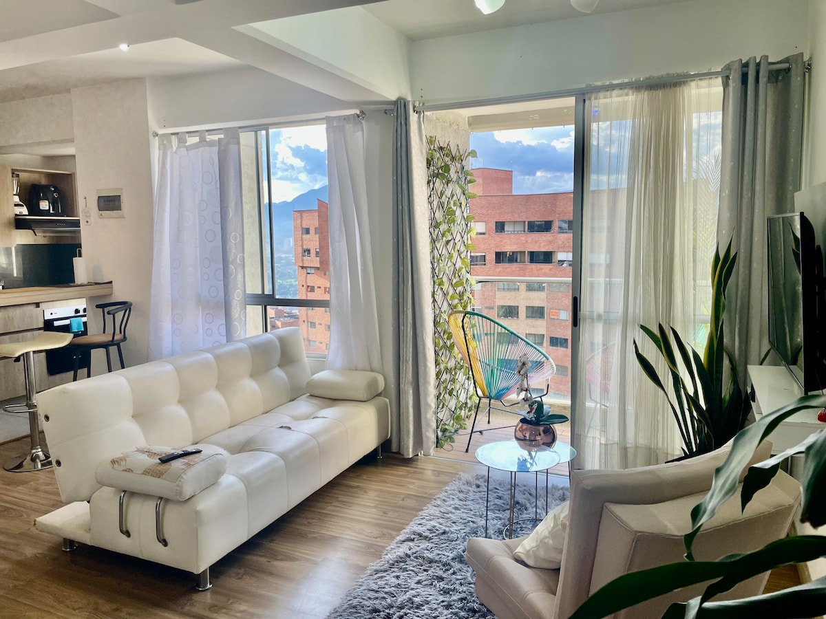 A modern living area is shown, featuring a white sectional sofa and large windows with sheer curtains. A cozy area rug lies on the wooden floor, while plants and a decorative table add a touch of greenery. Nearby, a dining space is visible, alongside hints of mountain views.