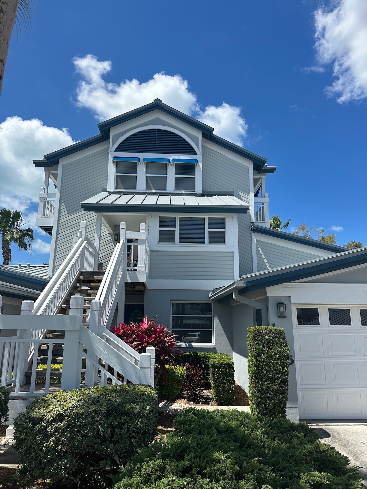 The exterior view of the townhome showcases a two-story structure with a peaked roof and balconies. Lush landscaping surrounds the entrance, while a set of stairs leads to the front door. Large windows allow plenty of natural light, offering a welcoming appearance.