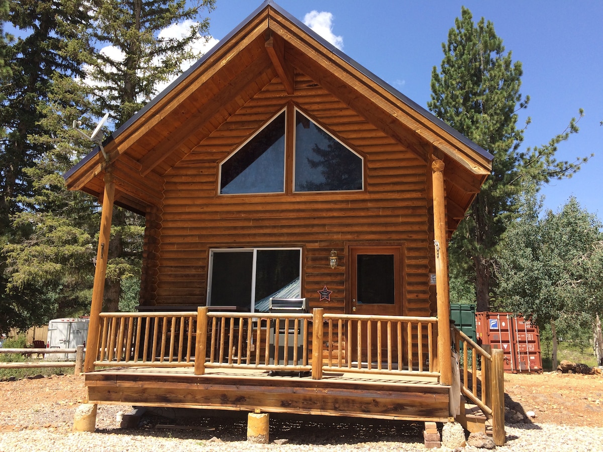 A rustic log cabin features a steeply pitched roof and large front windows, allowing natural light to fill the interior. A welcoming porch with wooden railings and a sturdy front step provides an inviting entrance. Surrounding trees offer a natural backdrop.