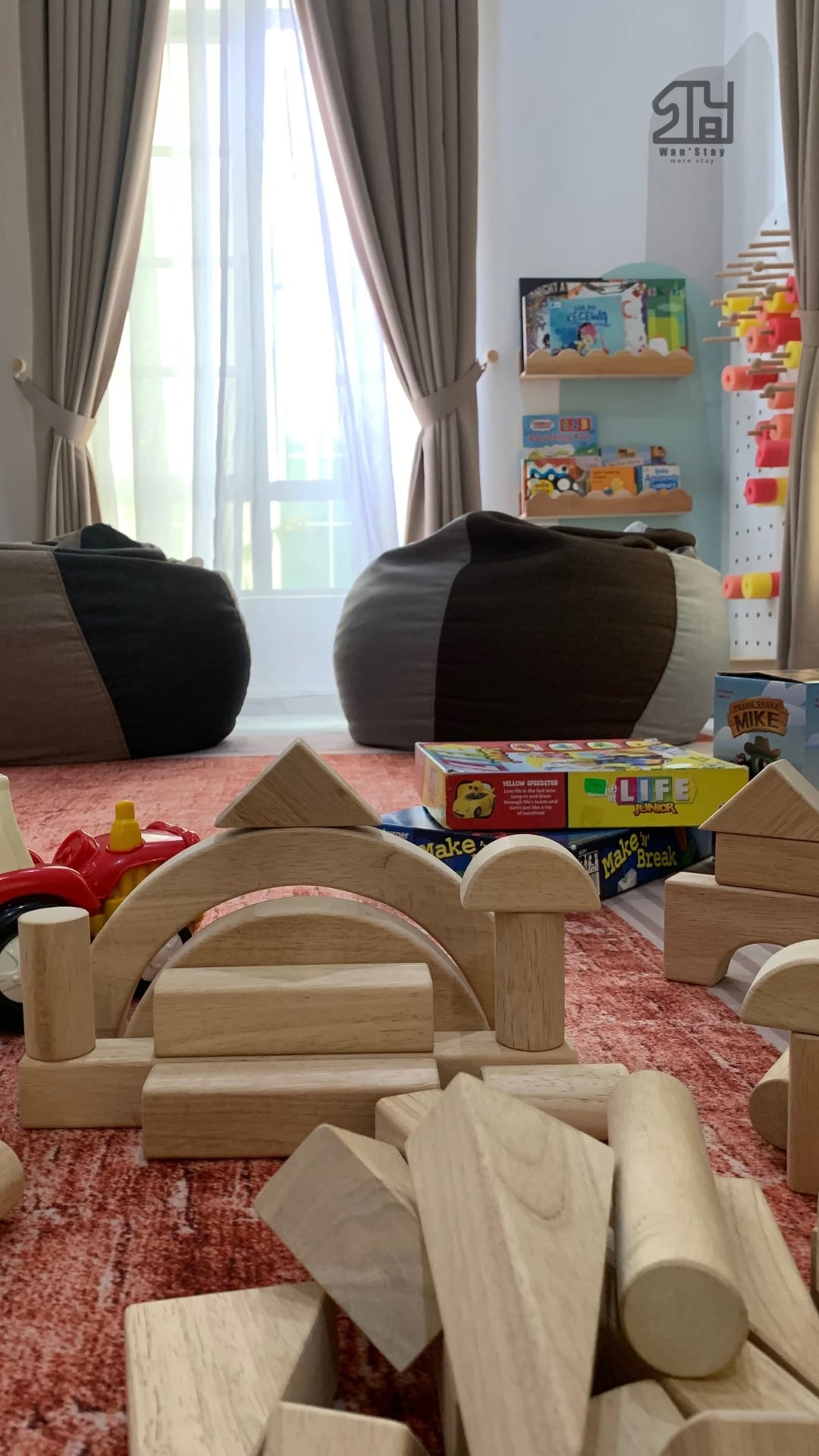 A play area is presented with soft bean bags, bright toys, and wooden building blocks. A colorful rug lies beneath, while natural light streams through large curtains, creating a cheerful atmosphere.