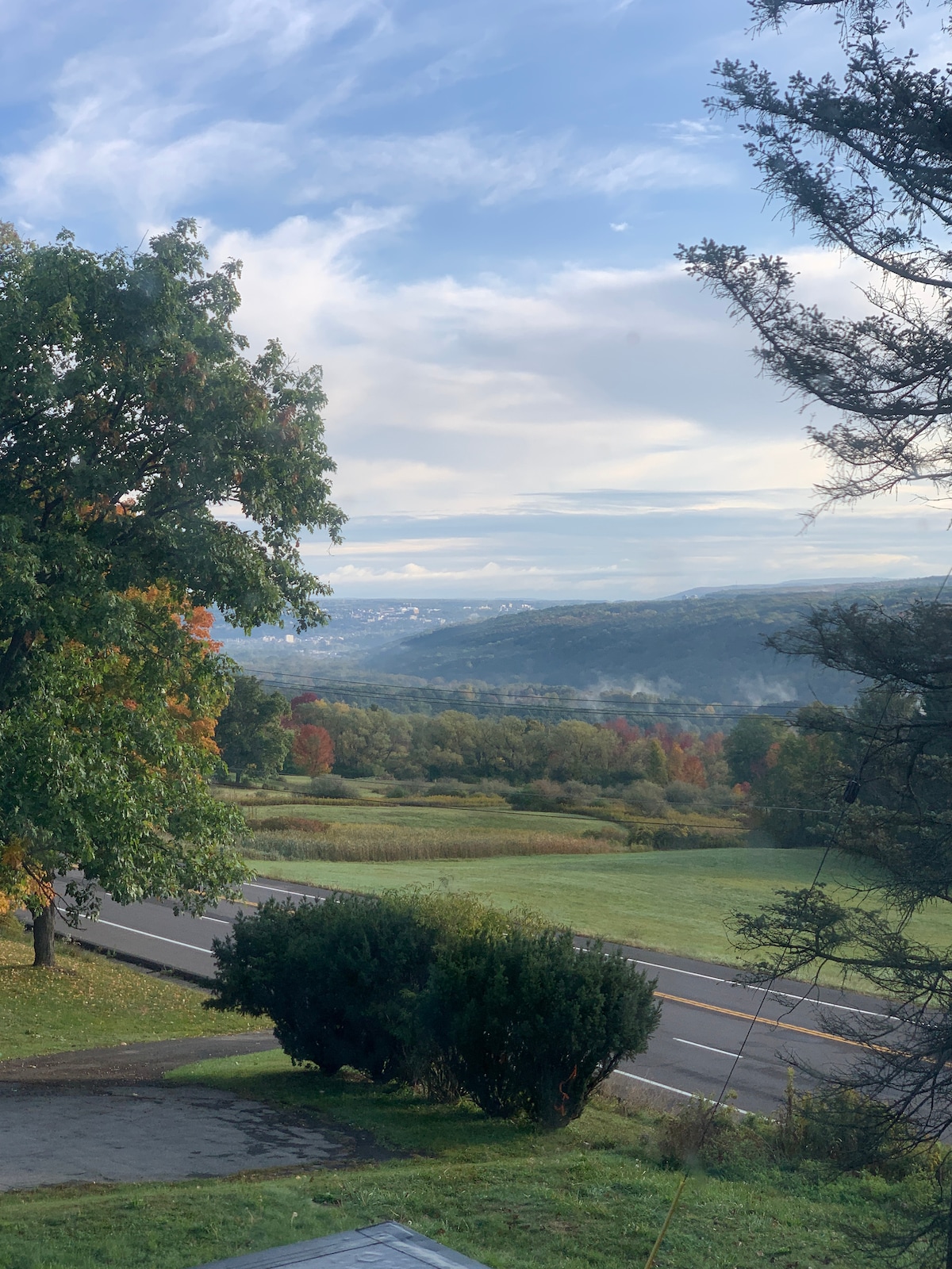A panoramic view of lush green hills and trees, with hints of autumn colors along the landscape. A winding road can be seen, leading through the valley, under a partly cloudy sky that stretches into the distance.