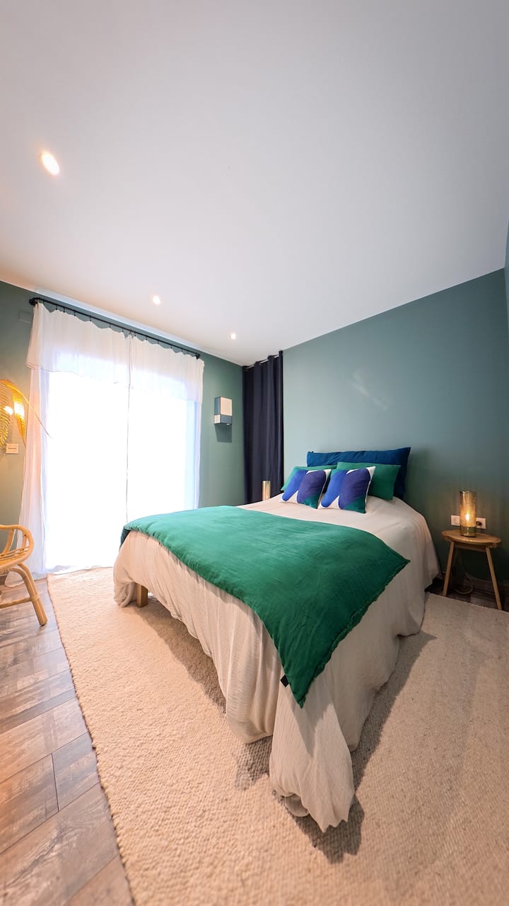 Camera luminosa e rilassante nei toni del verde acqua e del blu. Comodo letto matrimoniale con lenzuola curate, atmosfera accogliente e contemporanea. Ampia vetrata con veli, illuminazione soffusa, arredi naturali e tappeti chiari.