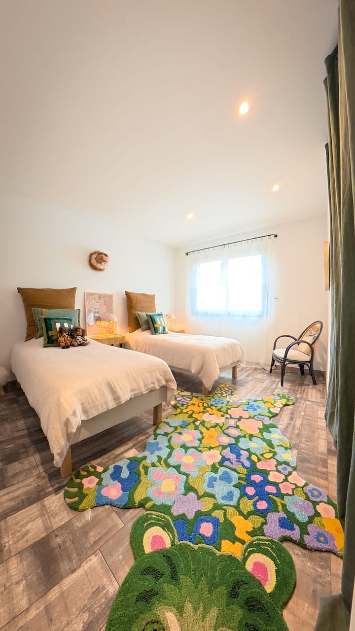 Camera bambini luminosa con due letti singoli, arredamento dolce e accogliente. Tappeto giocoso a forma di coccodrillo, colori allegri, poltrona accogliente e luce naturale.