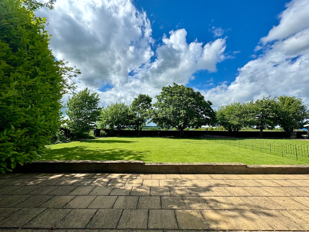 A well-maintained garden area is depicted, featuring a lush green lawn surrounded by trees under a partly cloudy sky. The stone pathway leads through the garden, offering a serene outdoor space for relaxation.