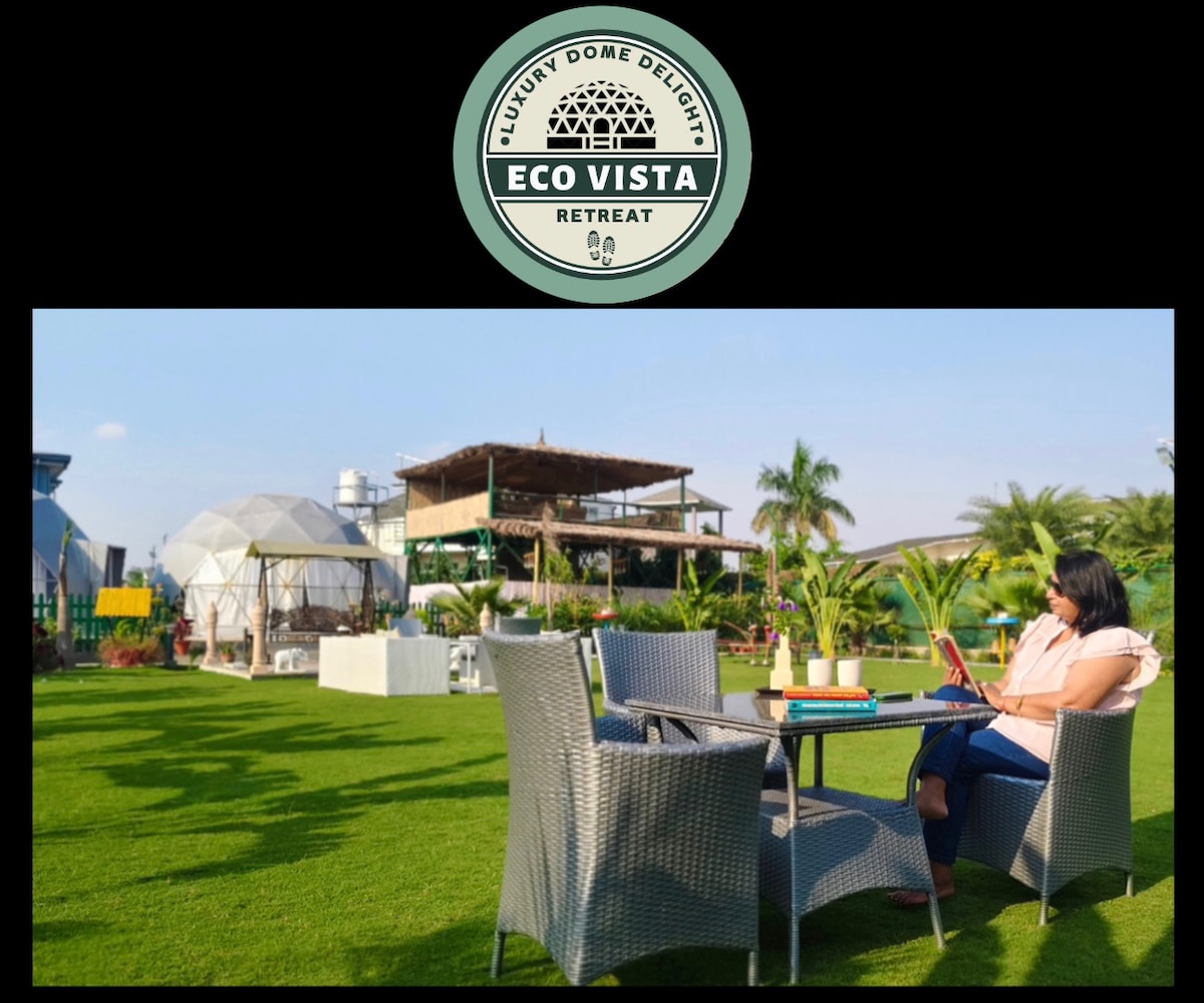 A green lawn is equipped with a round table and four chairs, providing a serene outdoor seating area. Nearby, luxury domes are visible, set against a clear sky, while palm trees and a machaan structure enhance the tranquil ambiance.