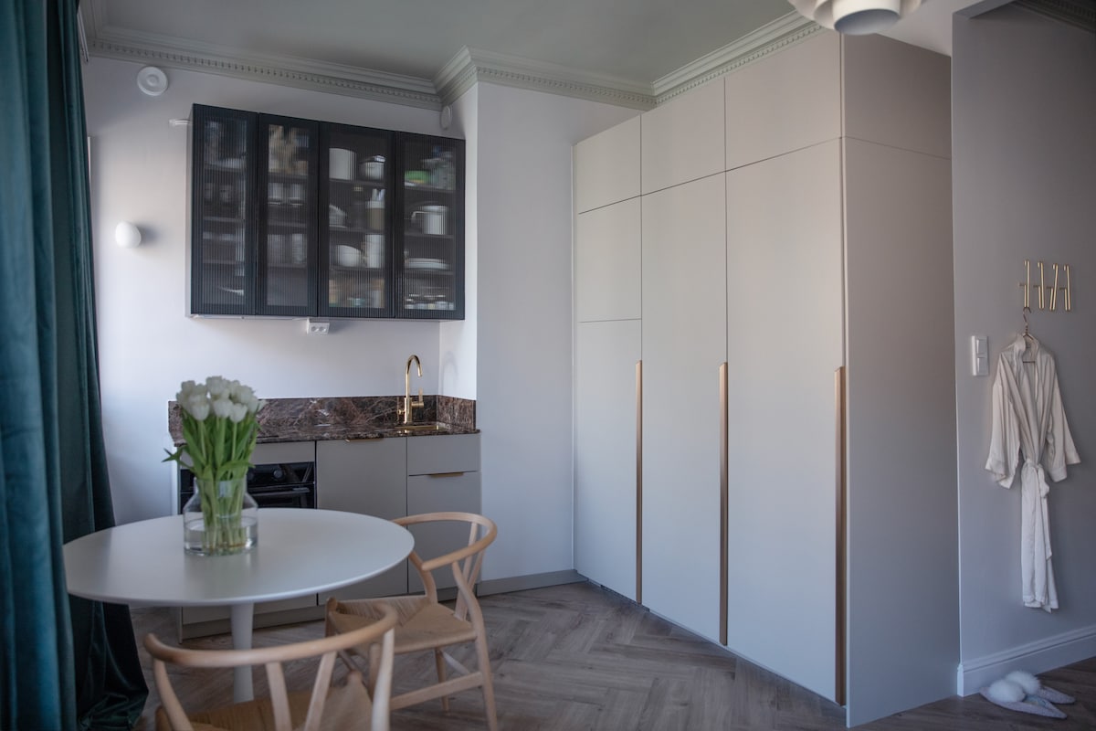 A modern kitchen area features sleek cabinetry with glass doors displaying dishware. Natural light highlights a round dining table with two wooden chairs. A stylish, wrapped robe hangs on the wall, complementing the innovative design of the space.