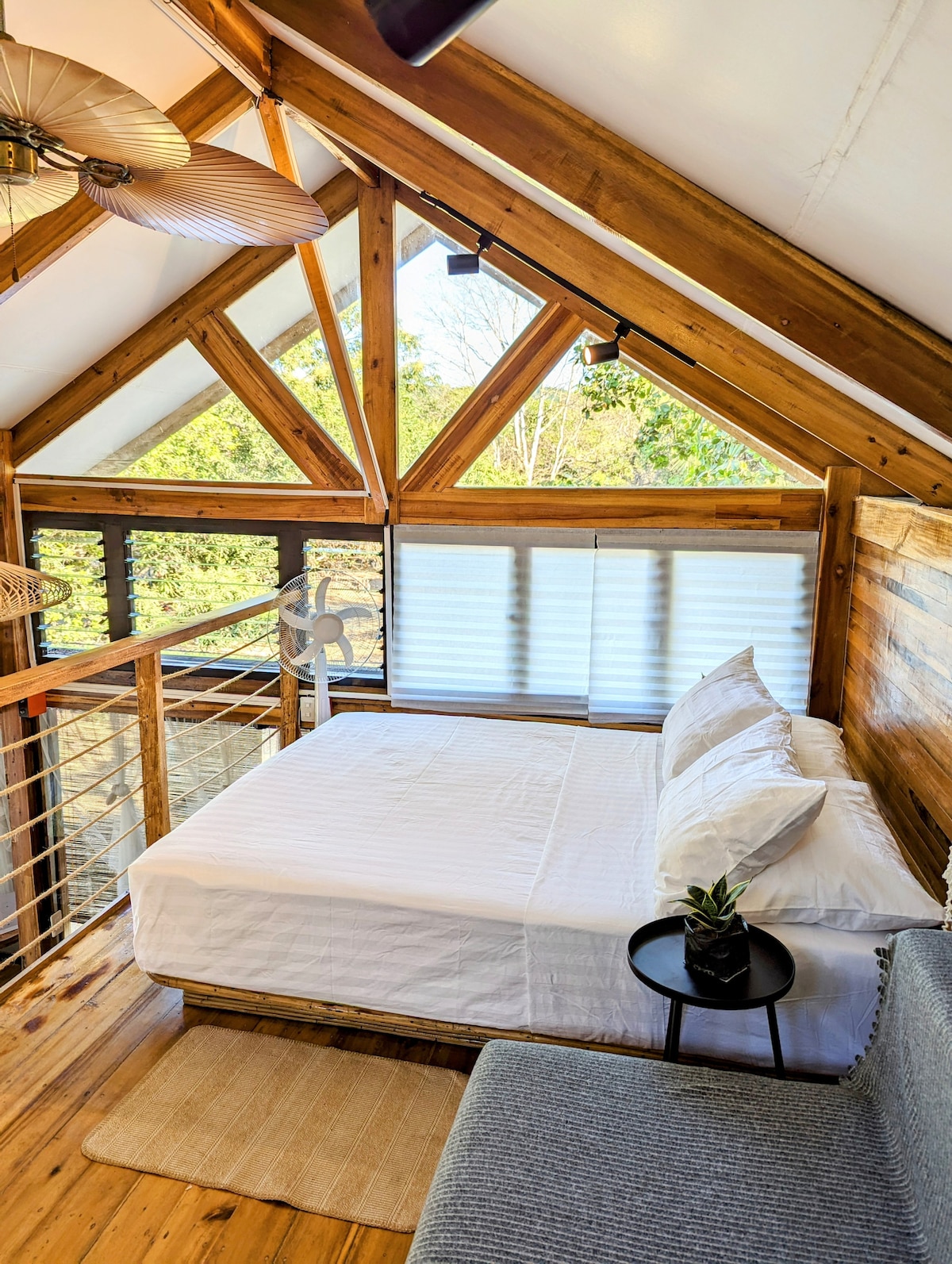 A loft bedroom features a spacious bed with white linens, set against wooden accents and high ceilings. Light filters through shaded windows, while a fan provides a gentle breeze. A small side table holds a decorative plant, enhancing the serene atmosphere.