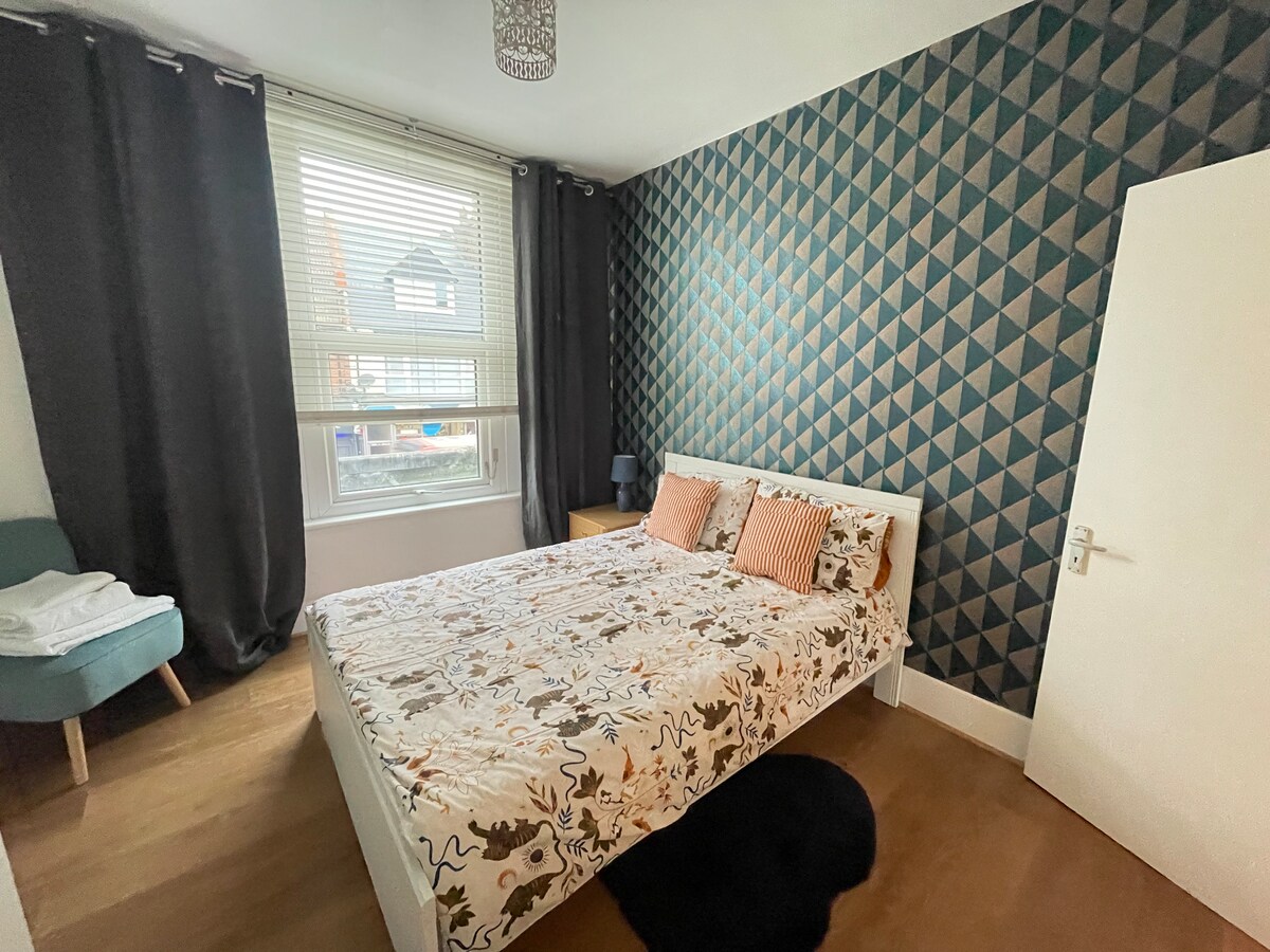 A modern bedroom features a double bed with a patterned bedding set and contrasting throw pillows. A large window with blinds allows natural light to fill the room, complemented by dark curtains. A small bedside table and a cozy chair are also present.