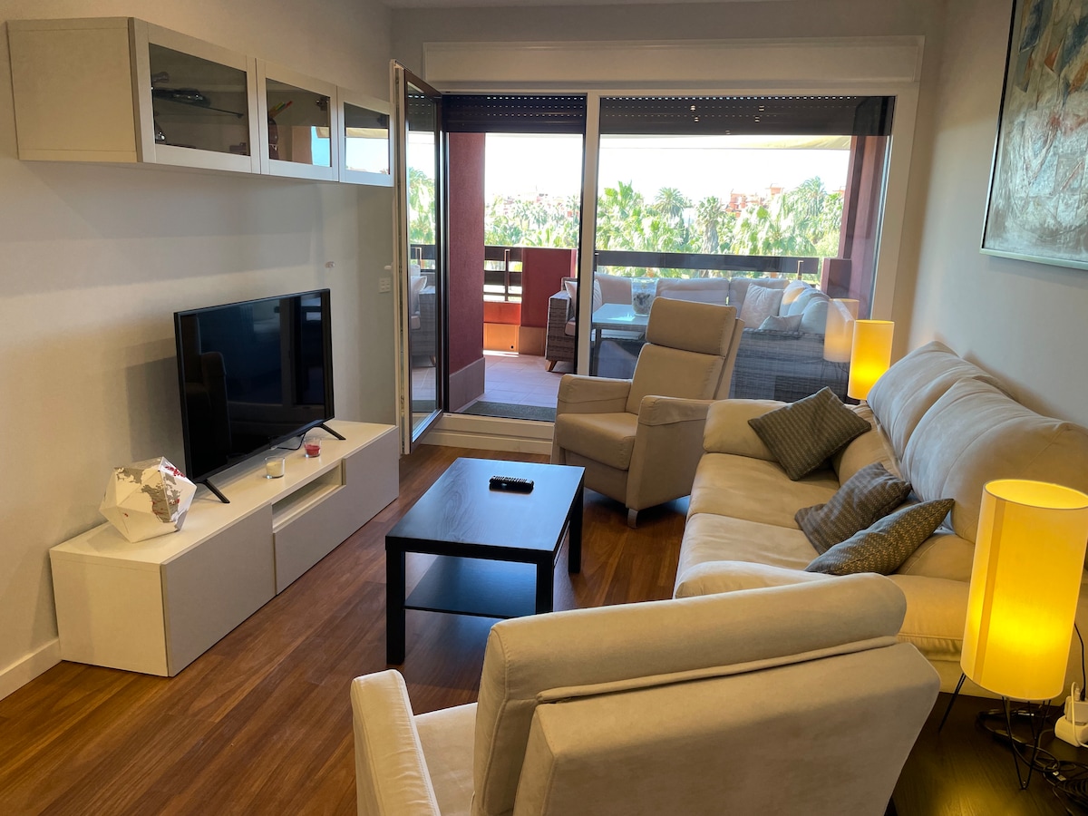 A living area features a comfortable sectional sofa and a modern coffee table, positioned near a television unit. Large glass doors open to a balcony, allowing natural light to fill the space. The warm wood flooring enhances the ambiance of the room.