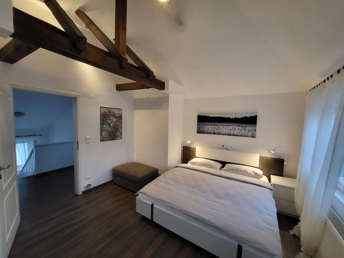 The spacious bedroom features a large bed with crisp linens and bedside lamps on either side. A minimalist nightstand complements the bed, and a comfortable seating area is visible on one side. Natural light filters in through a window, enhancing the calm ambiance of the room.