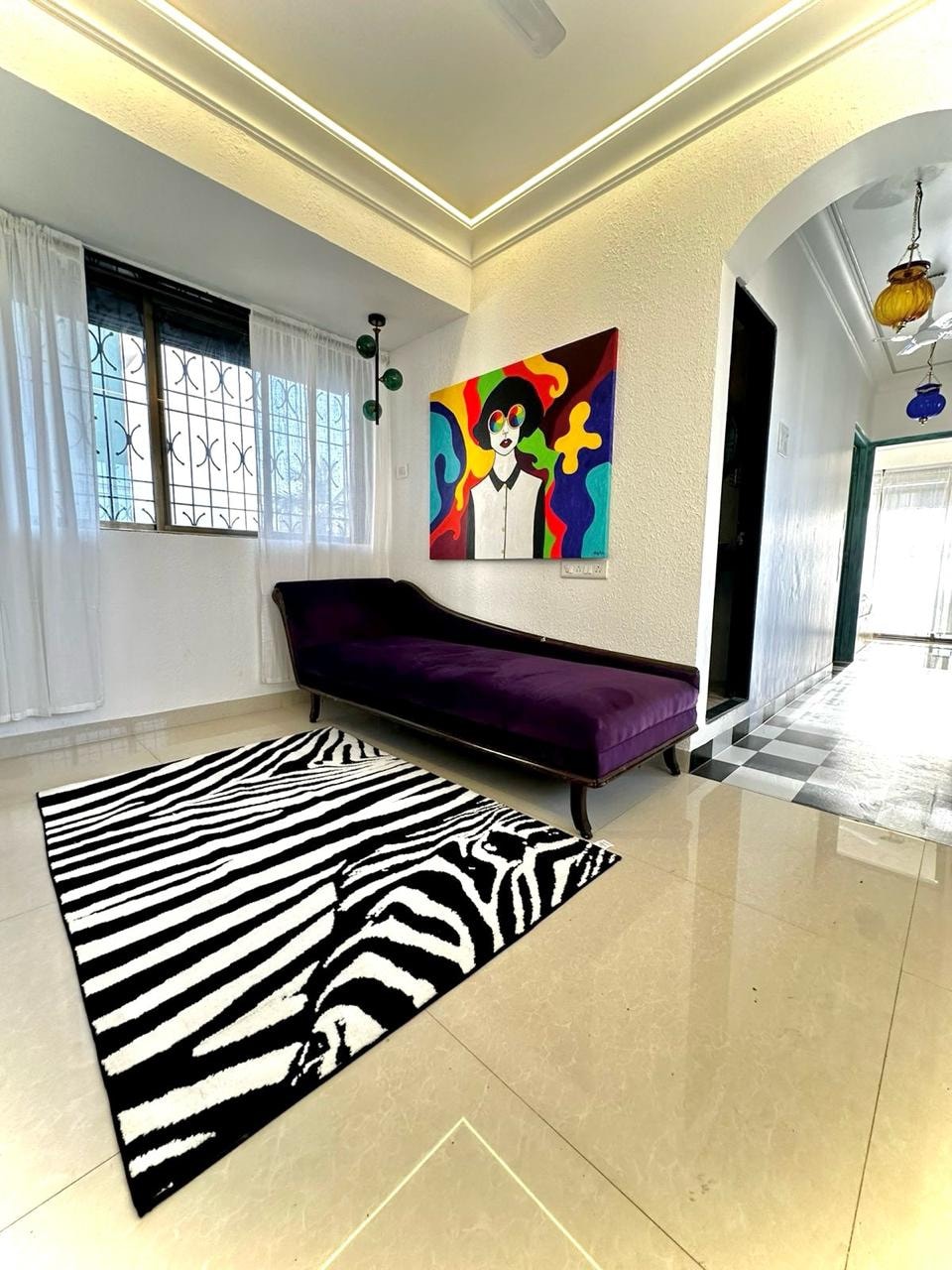 A cozy corner features a purple chaise lounge set against a wall adorned with a colorful abstract painting. A bold black and white zebra-striped rug adds texture to the glossy floor, while natural light filters through sheer white curtains hanging beside a window.