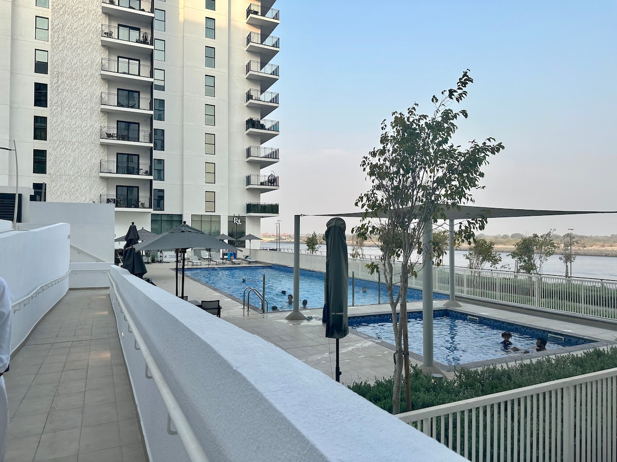 A view of a resort-style pool area, featuring a rectangular swimming pool with lounge chairs and umbrellas along the edges. The surrounding green plants provide a natural touch, while the multi-story building stands prominently nearby, reflecting a calm waterfront atmosphere.