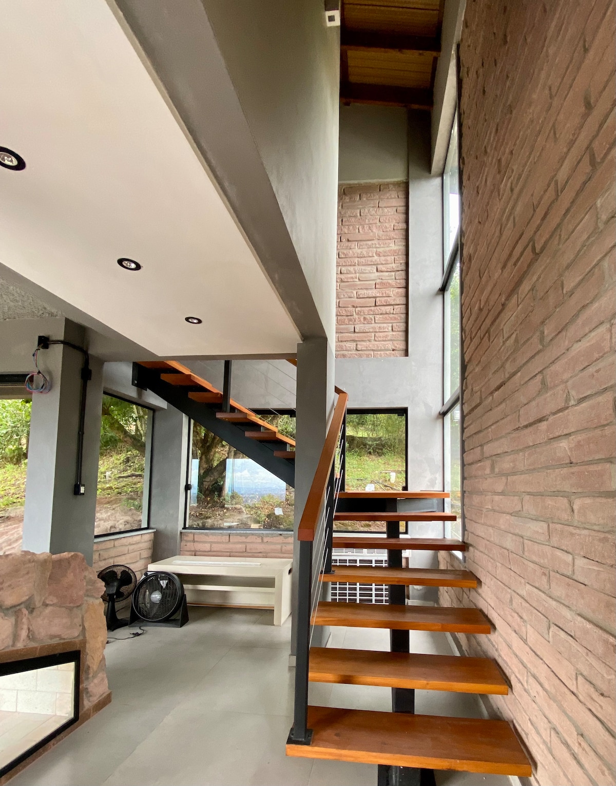 A staircase with wooden steps is positioned against a stone wall, leading to an upper level. Large windows allow natural light to flood in, providing a view of the greenery outside. The open layout creates a sense of spaciousness.