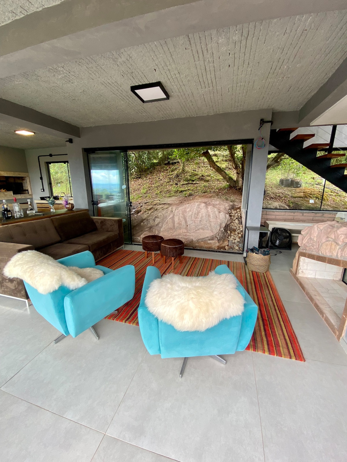 A comfortable seating area features two bright blue chairs with soft white throws, positioned around a pair of wooden stools. A textured rug adds warmth to the space, while large glass doors provide views of the natural surroundings.