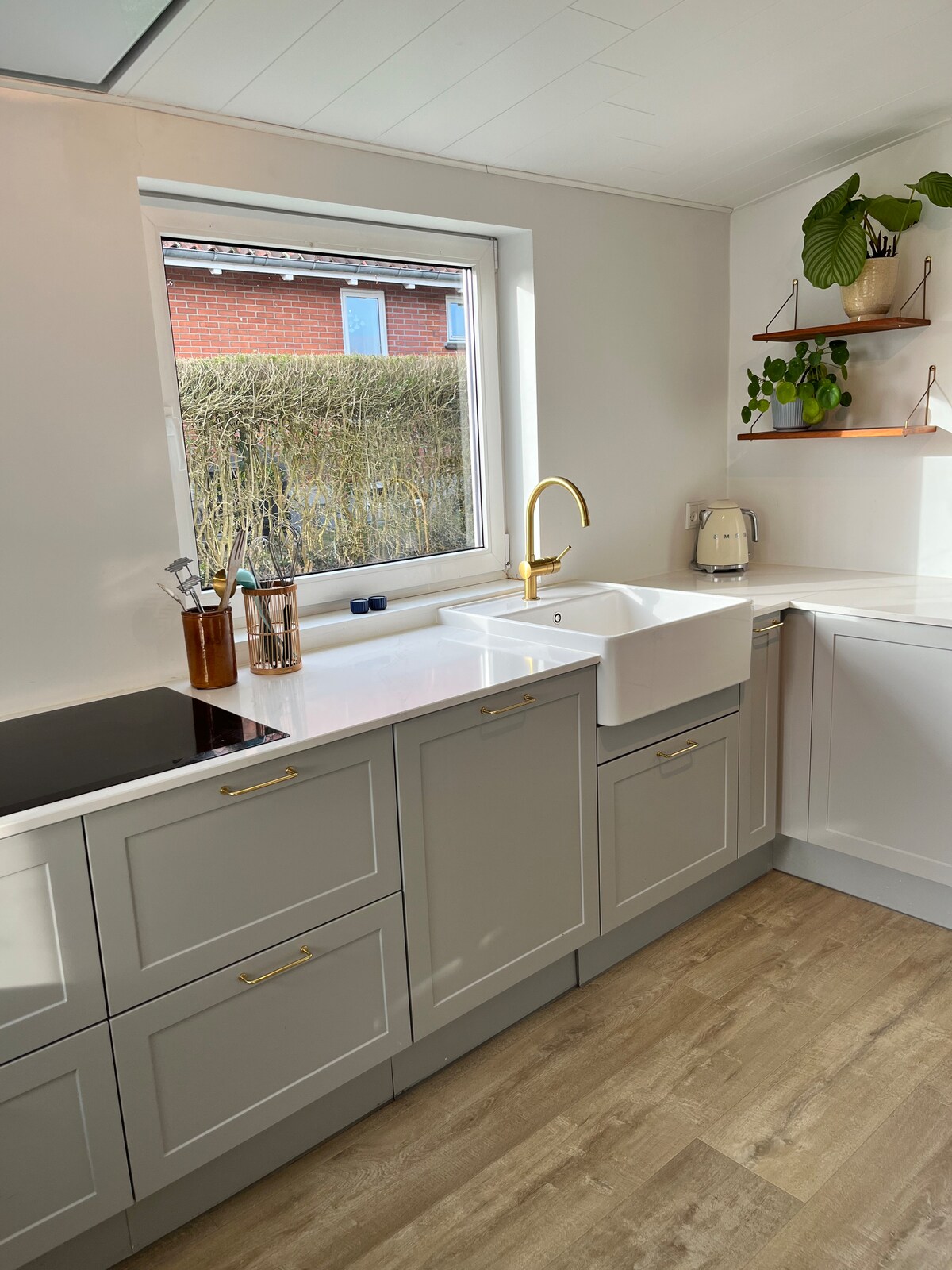 A modern kitchen features a large window overlooking greenery, allowing natural light to brighten the space. A white sink is paired with a gold faucet, complemented by sleek cabinetry with gold handles. Open shelving displays attractive plants and kitchen essentials.