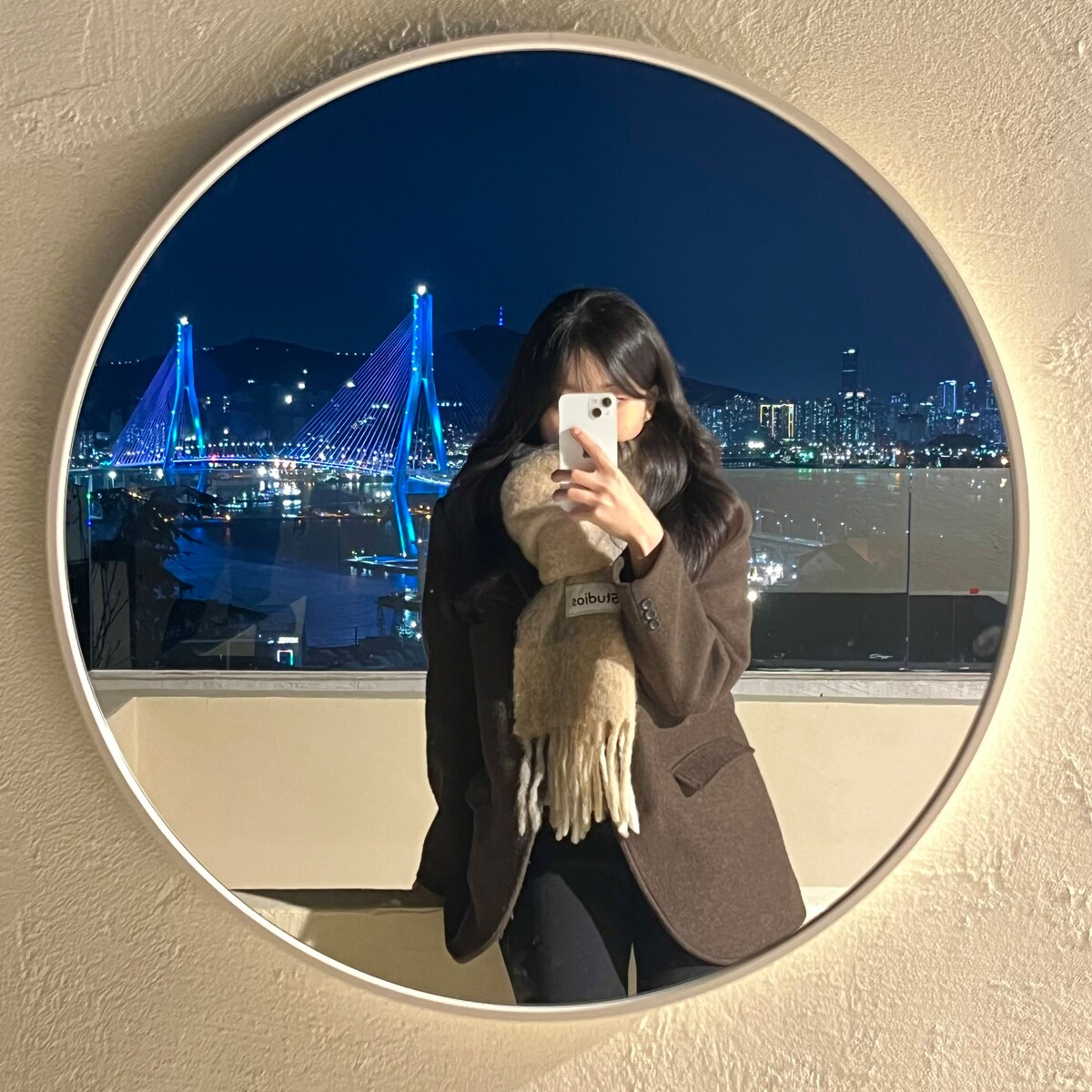 A round mirror reflects a person standing on a balcony, overlooking the brightly illuminated city skyline at night. The colorful lights of the bridge are visible in the background, enhancing the scene's urban ambiance.