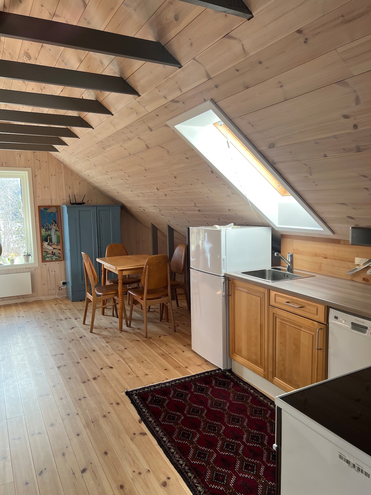 A bright and airy space features sloped wooden ceilings and warm-toned walls. A wooden dining table with chairs is positioned near a window, complemented by a dark cabinet. Kitchen amenities, including a refrigerator and sink, are visible against the wall, enhancing the functional design.