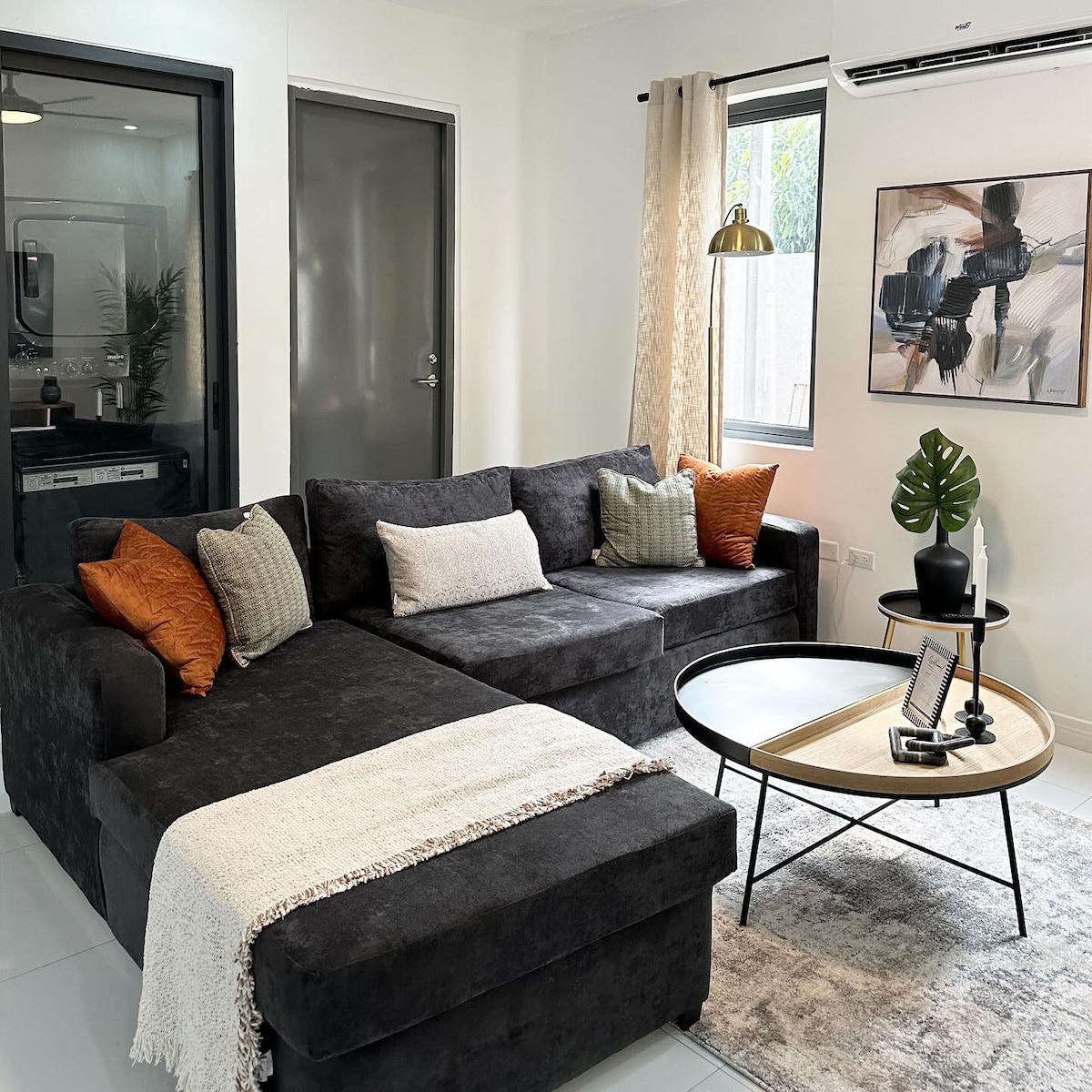 A modern living area features a cozy sectional sofa in dark fabric, accented by decorative pillows. A round coffee table occupies the center, complemented by a light area rug. Natural light filters in through the windows, enhancing the space's inviting feel.