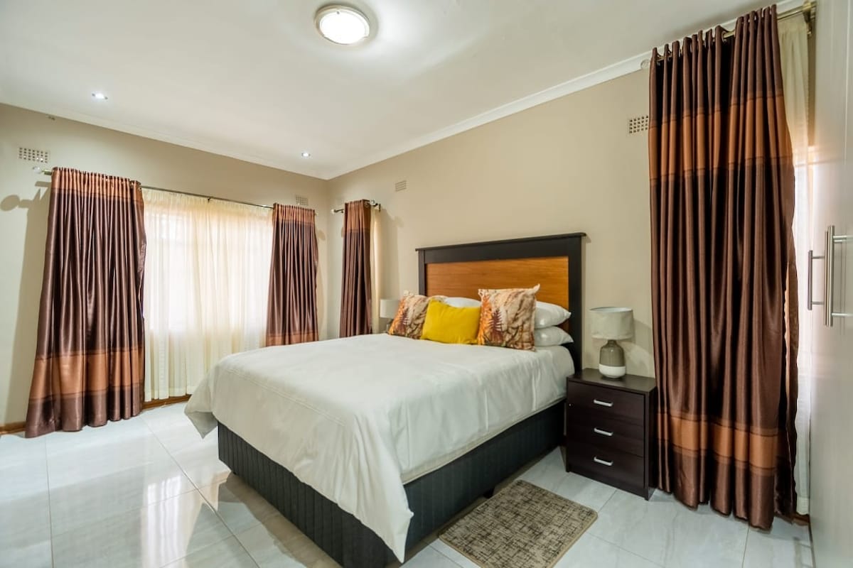 A spacious bedroom features a large bed dressed in neutral linens, accented by decorative pillows. Light flows in from two windows, framed by elegant curtains. A dark wooden bedside table stands beside the bed, enhancing the room's serene ambiance.