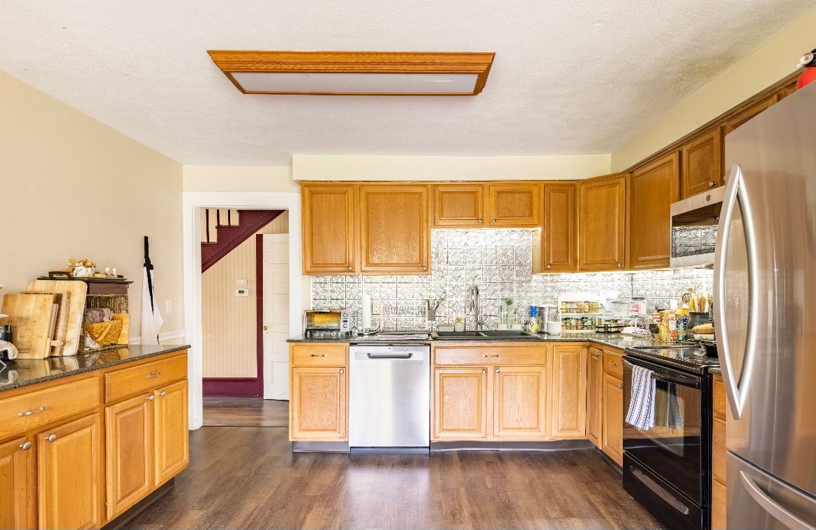 A well-equipped kitchen features wooden cabinetry with granite countertops. Stainless steel appliances, including a dishwasher and a stove, complement the space. A textured backsplash adds a modern touch, while natural light enters through a window, creating an inviting atmosphere for cooking and gathering.