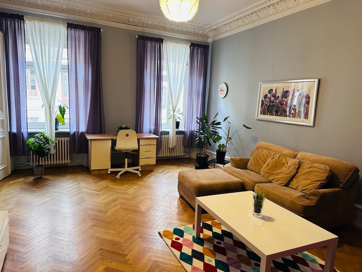 A spacious living area features large windows dressed in sheer curtains, allowing natural light to brighten the room. A comfortable sofa and a neutral coffee table are complemented by a colorful area rug. A desk and several potted plants add to the inviting ambiance.