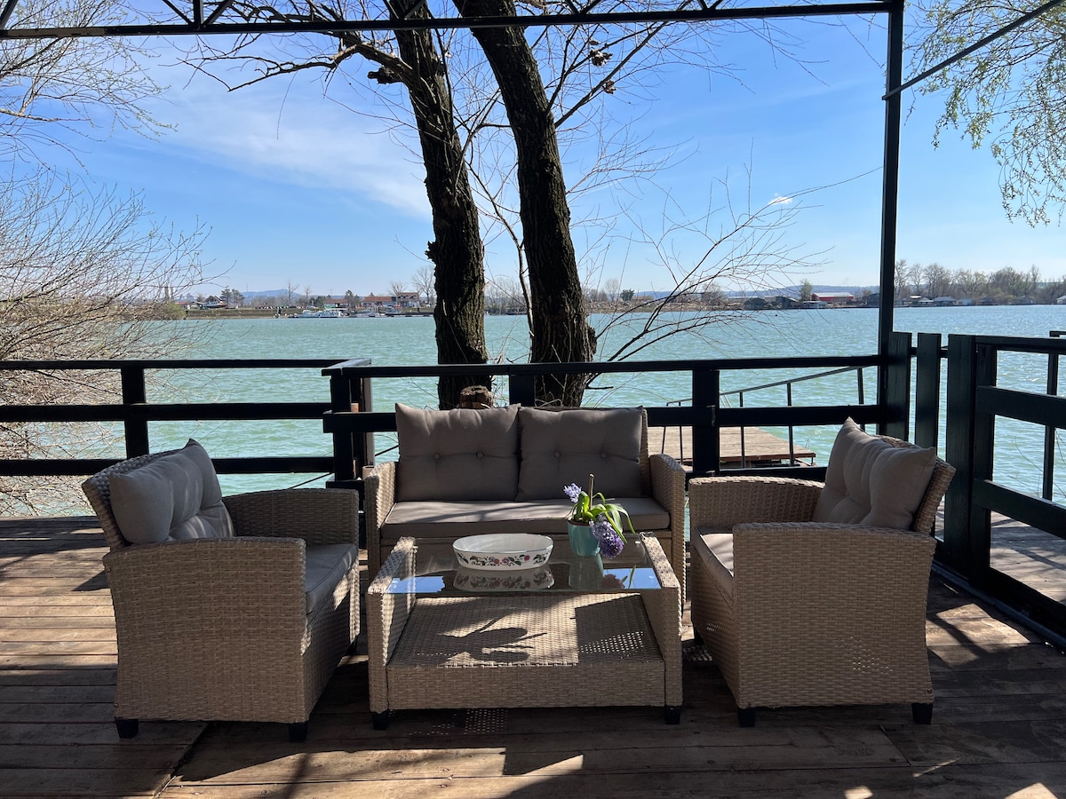 A comfortable patio area features a glass-top table surrounded by three plush seating arrangements, providing a relaxing outdoor space. The serene view of the river is framed by nearby trees, creating a peaceful environment for guests to unwind.