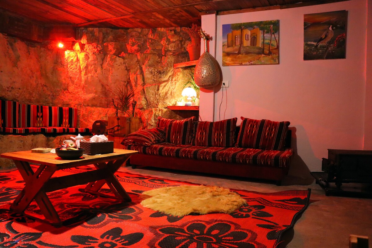 A cozy living area features a vibrant patterned rug and a wooden table at the center. Plush seating is arranged against a textured stone wall, complemented by warm red lighting. Artwork adorns the walls, adding a touch of character to the space.