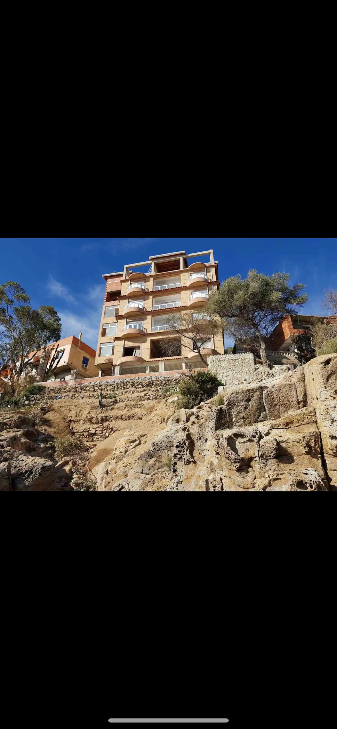 A multi-story building is situated on a rocky hillside, showcasing multiple balconies with views of the surroundings. Natural vegetation and rocky terrain are visible in the foreground, enhancing the connection to the natural landscape.