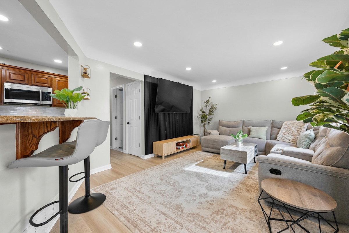 An open-concept living area features a sectional sofa and a modern entertainment unit with a wall-mounted TV. Natural light fills the space, complemented by a decorative area rug. A breakfast bar with gray stools offers additional seating, enhancing the inviting atmosphere.