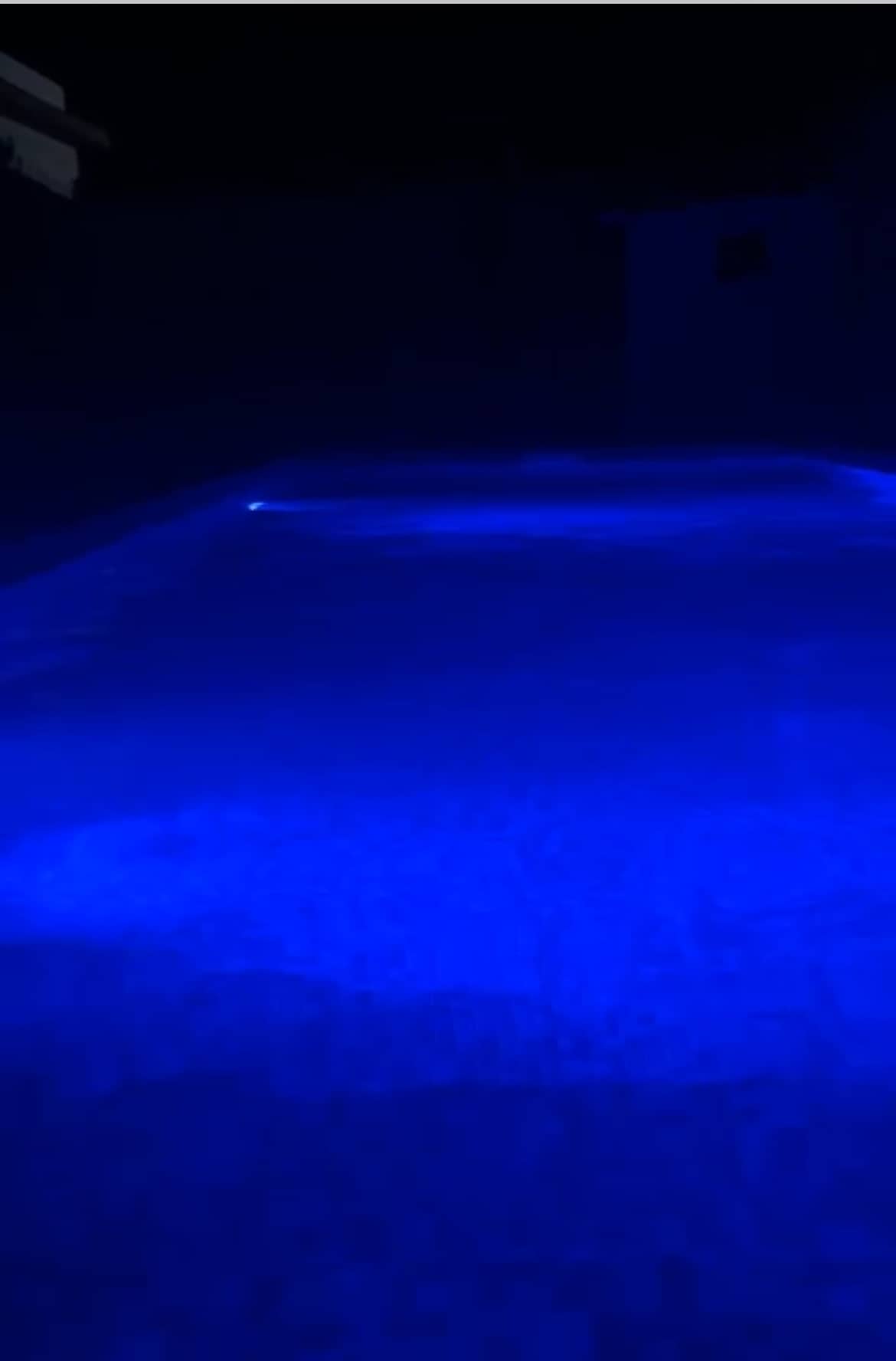 A spacious swimming pool emits a calming blue glow, highlighting the water's smooth surface in the evening light. The surrounding area is dimly lit, emphasizing the tranquil ambiance created by the illuminated pool.