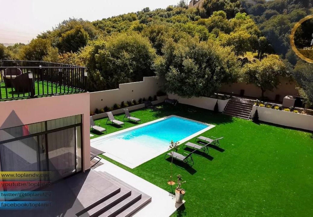 An outdoor area features a swimming pool surrounded by green grass and lounge chairs. The space is framed by lush trees and landscaping, creating a natural ambiance. A patio area adjacent to the pool is visible, offering additional seating options.
