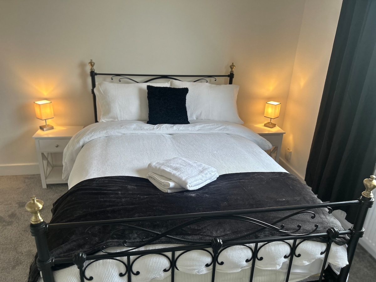 A cozy bedroom features a metal frame bed dressed in white linens, accented by a dark throw blanket. Two bedside tables with lamps provide soft illumination, and a plush towel is neatly arranged on the bed. Dark curtains frame a bright corner by the window.