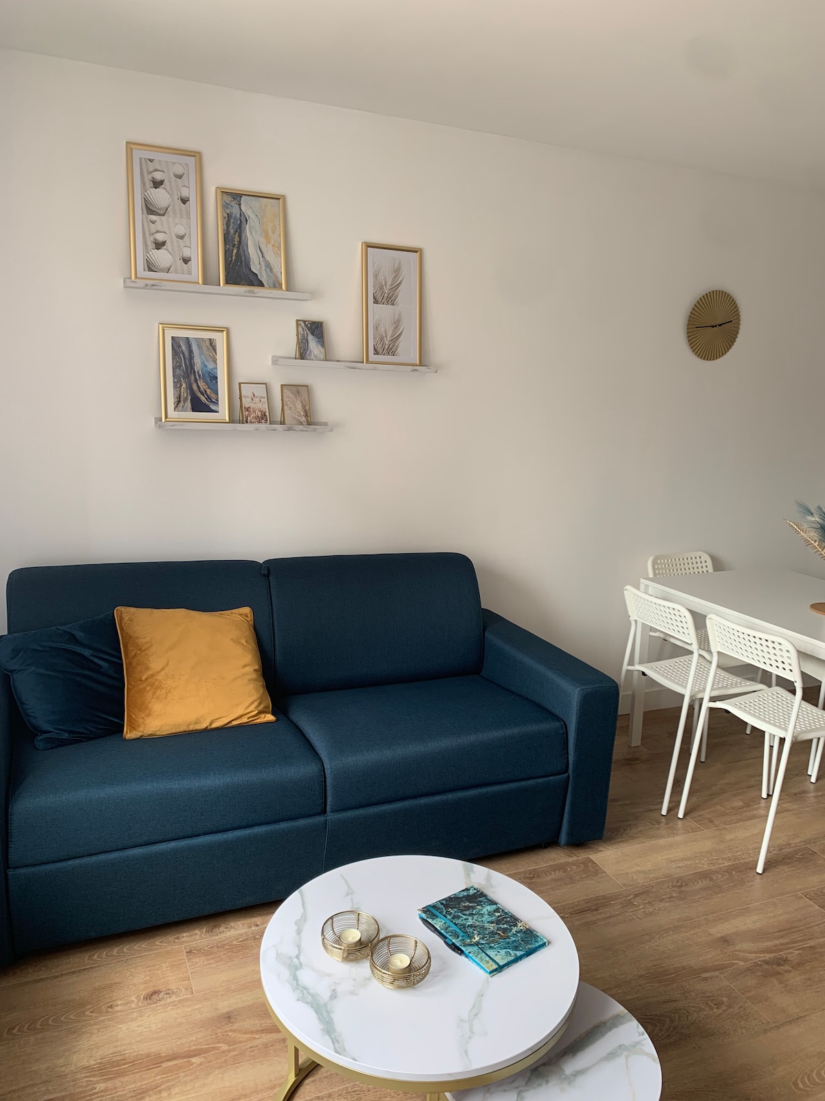 A modern sitting area features a dark blue sofa with a mustard pillow. Above, a set of framed artworks is displayed on white shelves. A round marble coffee table with two decorative glass candles sits in front of the sofa. A dining table with white chairs is positioned nearby.
