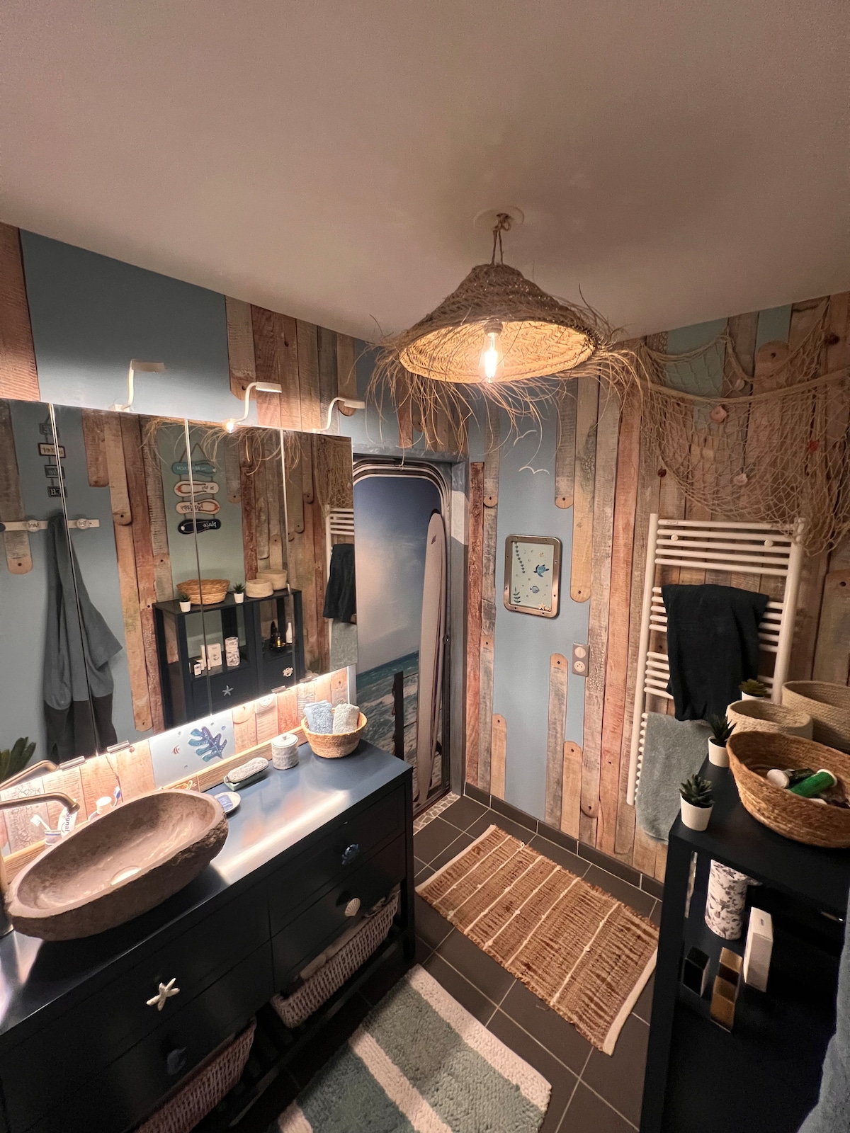 A spacious bathroom features wood-paneled walls adorned with nautical decor. A large mirror sits above a dark cabinet, reflecting the light from a woven pendant fixture. A small rug lies on the dark floor, complemented by decorative baskets and towels, creating a beach-inspired ambiance.