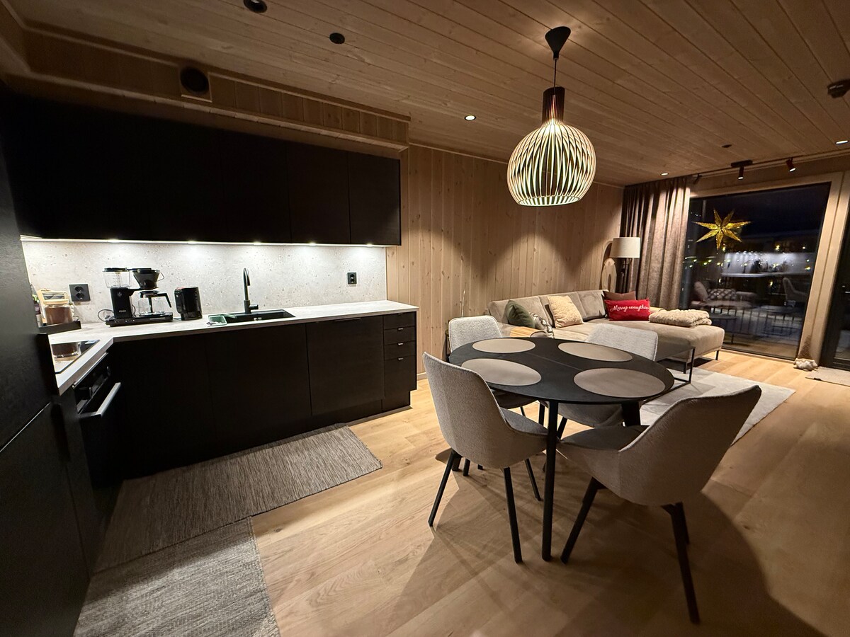 An open living area is featured, showcasing a modern kitchen with dark cabinetry and a light countertop. A round black dining table with four light-colored chairs is positioned beside the kitchen. A cozy seating area with a sofa is visible, and a balcony can be seen through the large windows.