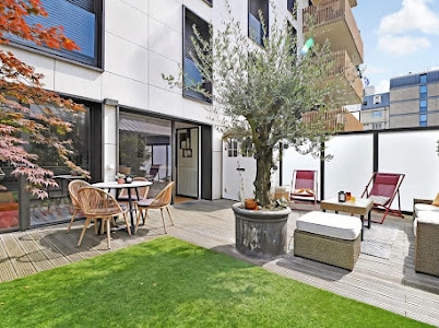 The outdoor terrace is furnished with a table and four chairs, alongside a seating area with two lounge chairs. A large potted olive tree serves as a focal point, with greenery covering the ground and a low glass barrier for privacy.